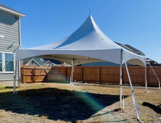 A white frame tent set up in a backyard with a wooden fence and house in the background under a clear blue sky.