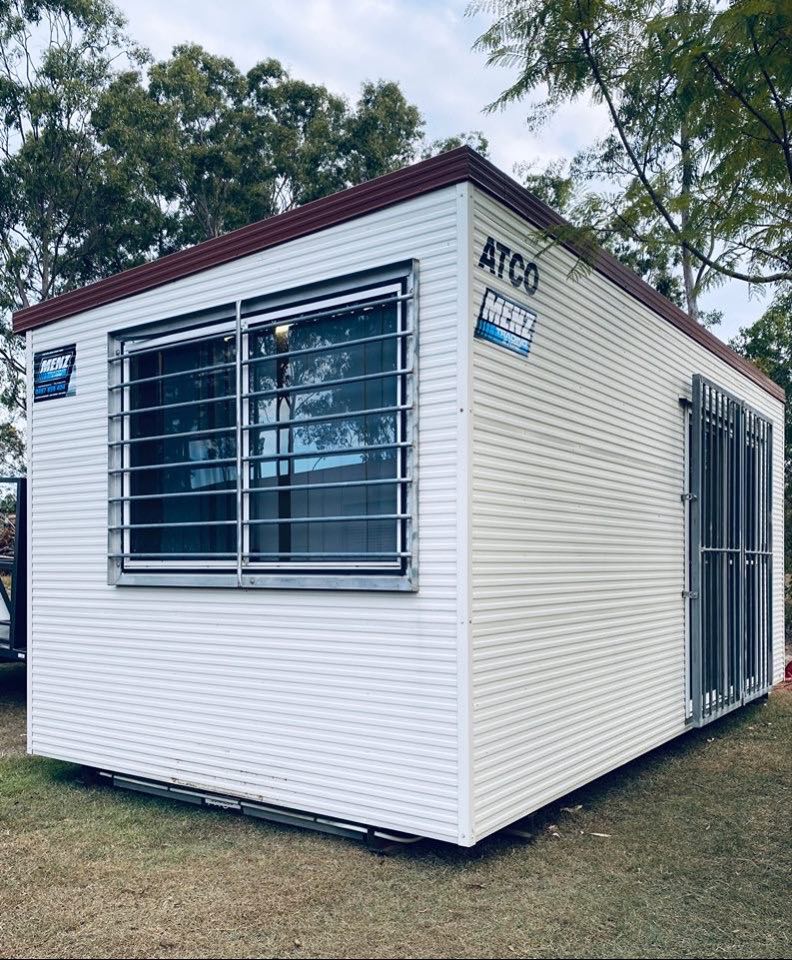 Small white portable office cabin with barred windows and a maroon roof on a grassy lot
