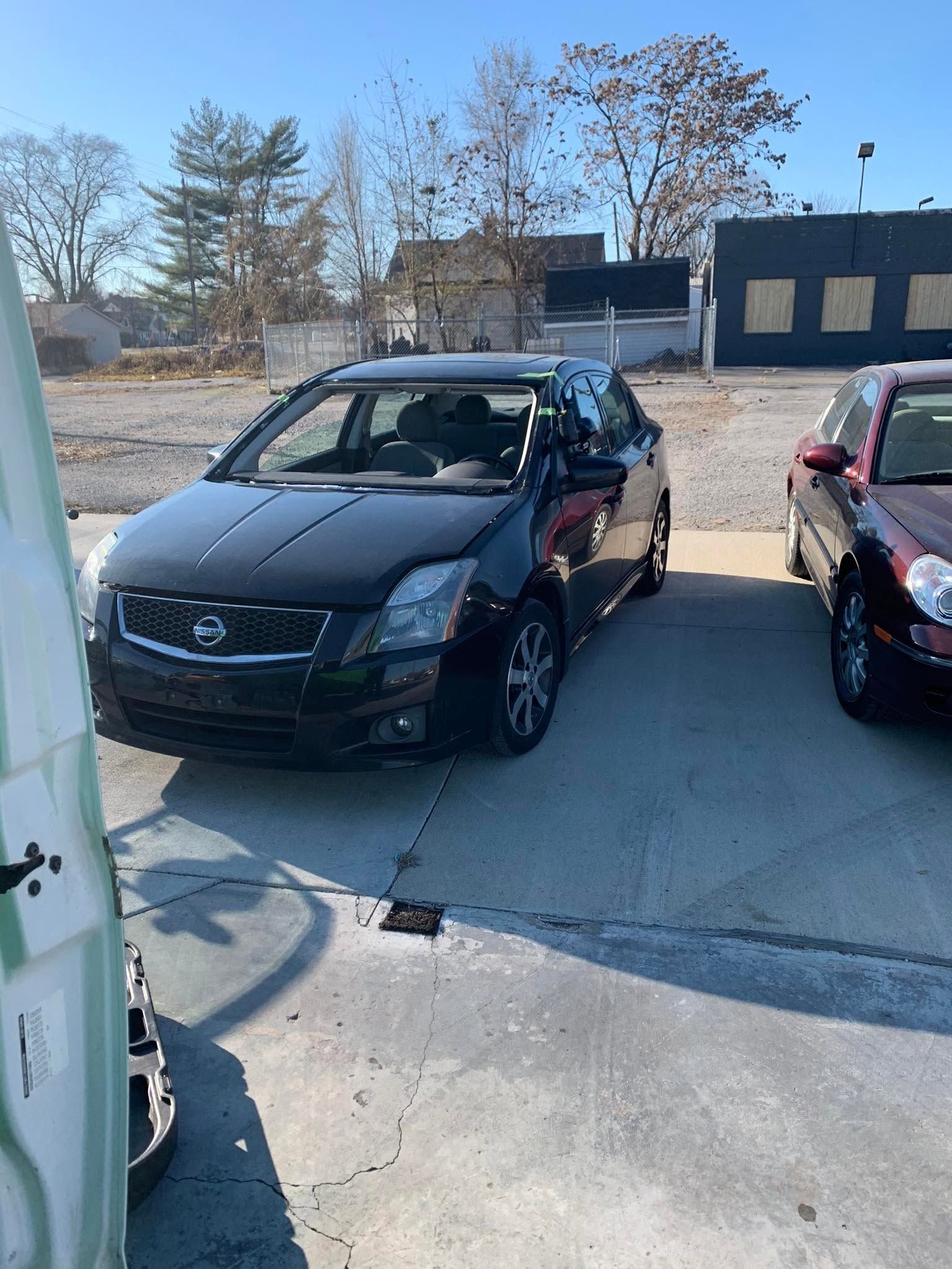 Black sedan parked in a sunny lot beside another car, with a person visible inside.