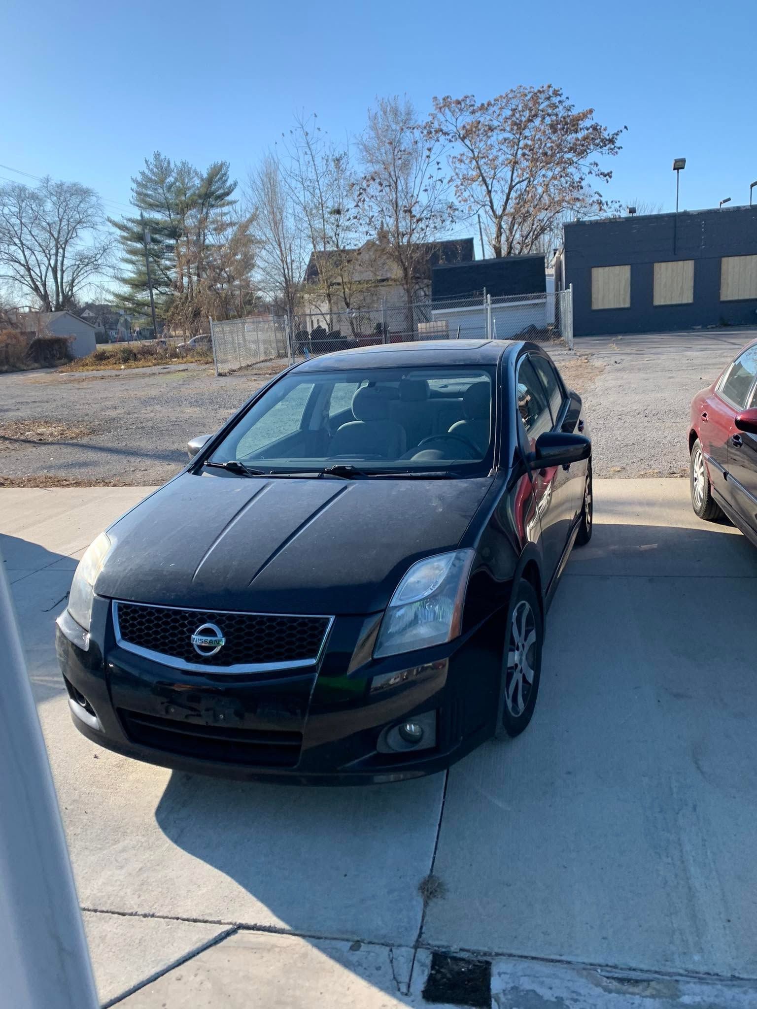 Black Nissan sedan parked in a driveway, with a red car beside it and a building in the background.