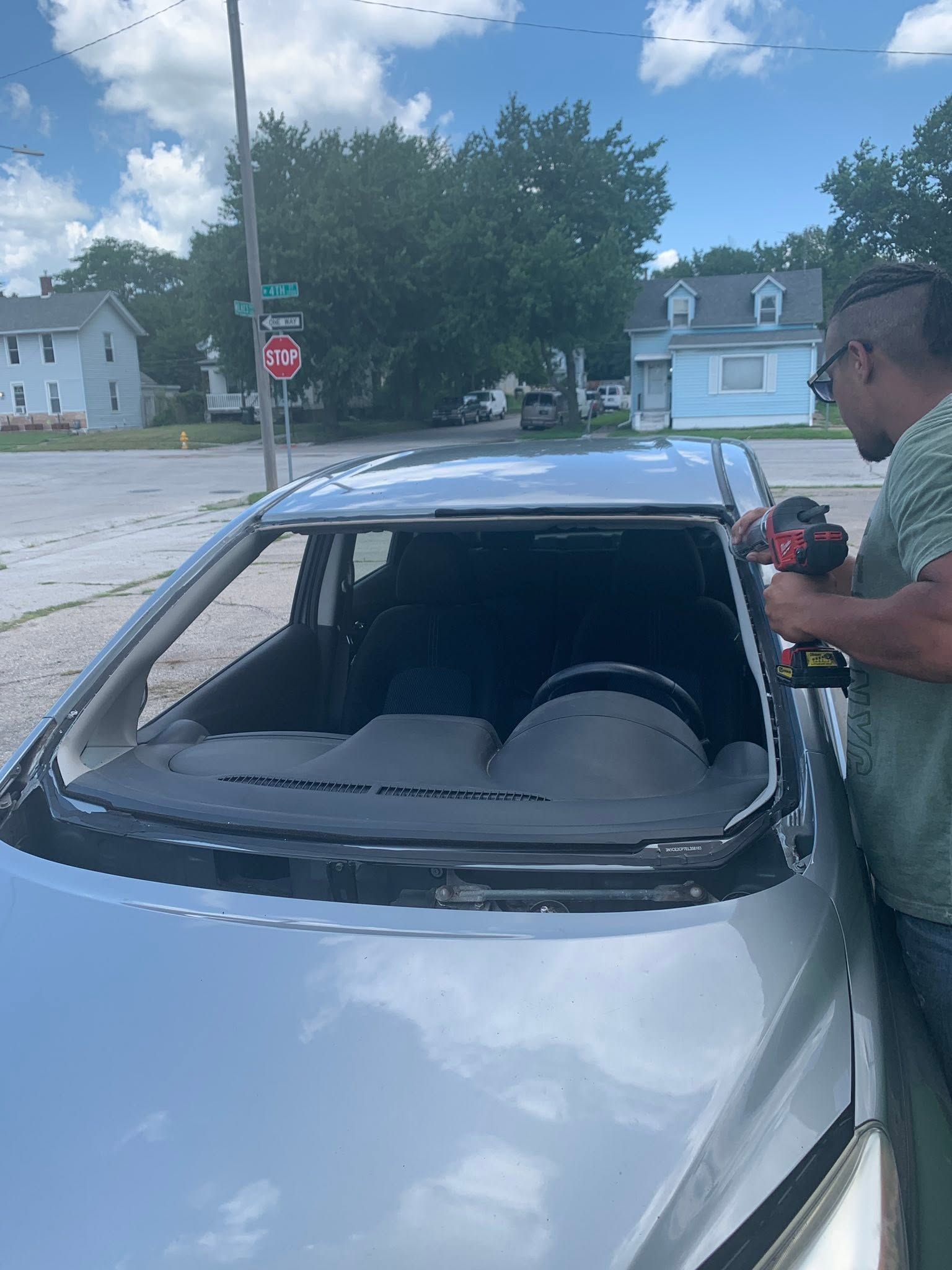 Silver car with shattered rear window on a street, with a person standing beside it holding a tool.