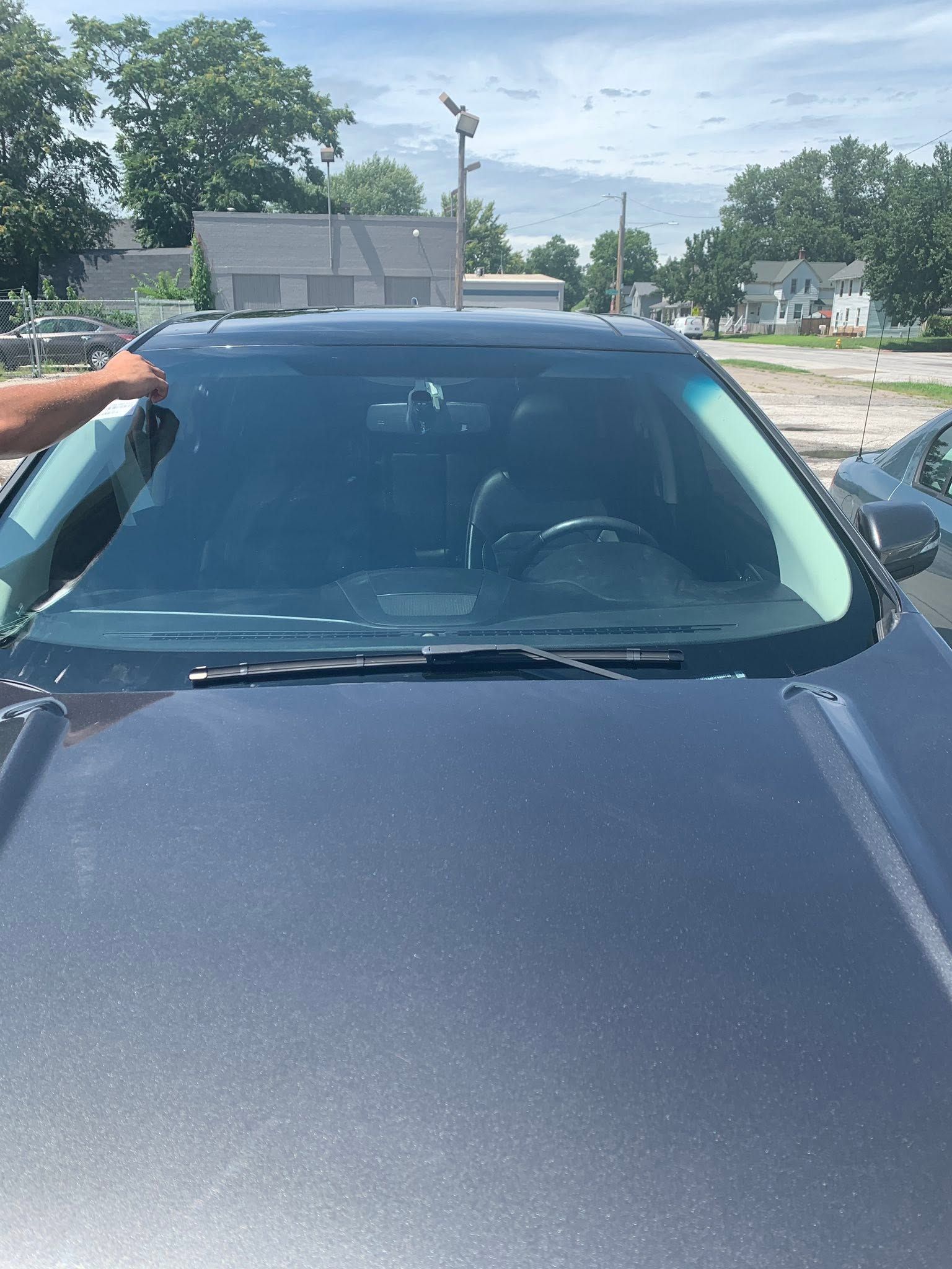 Hand on black car hood in a sunny outdoor parking lot, with trees and buildings in the background