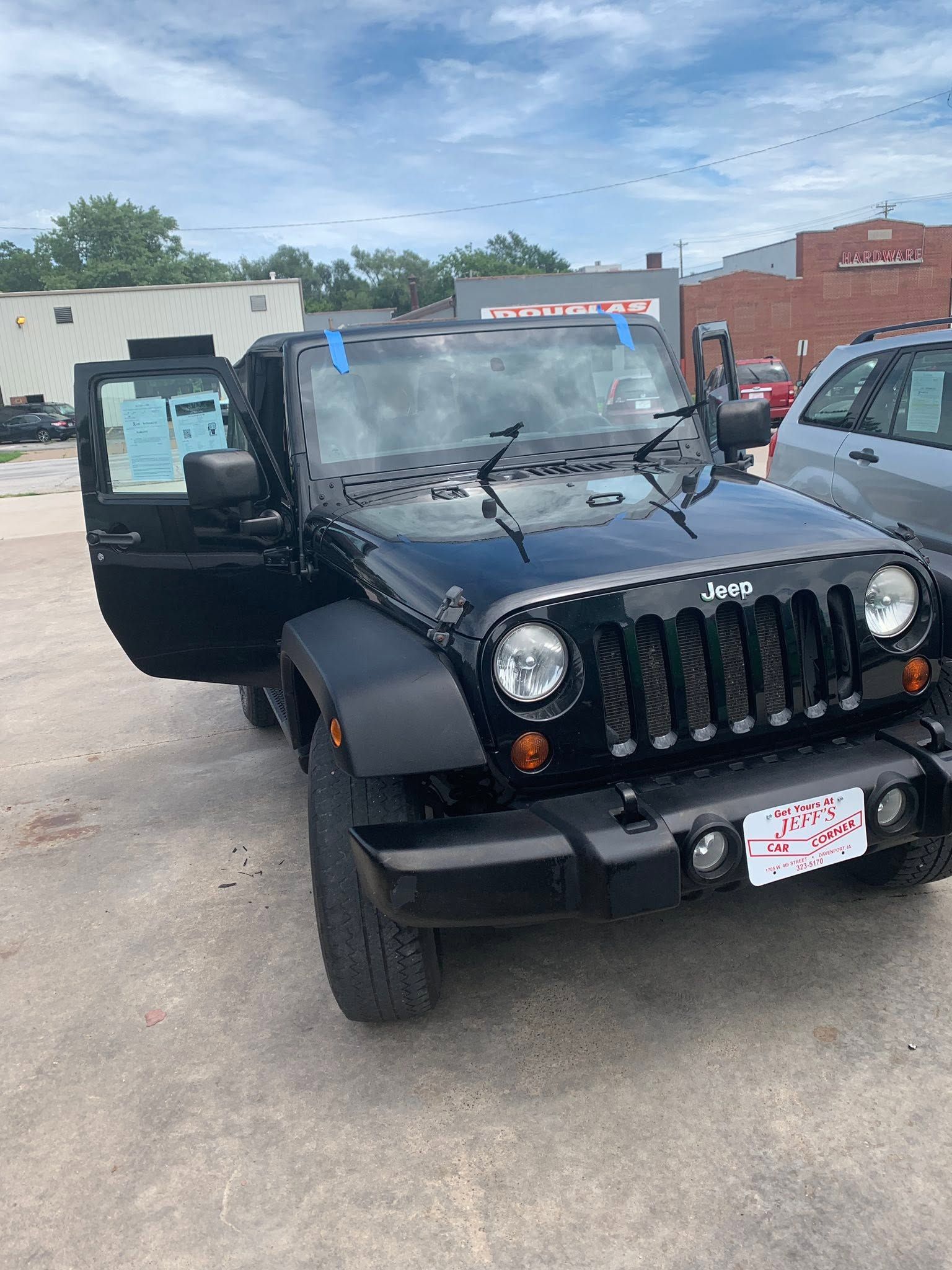 Black Jeep Wrangler with driver door open, parked outdoors in a lot.