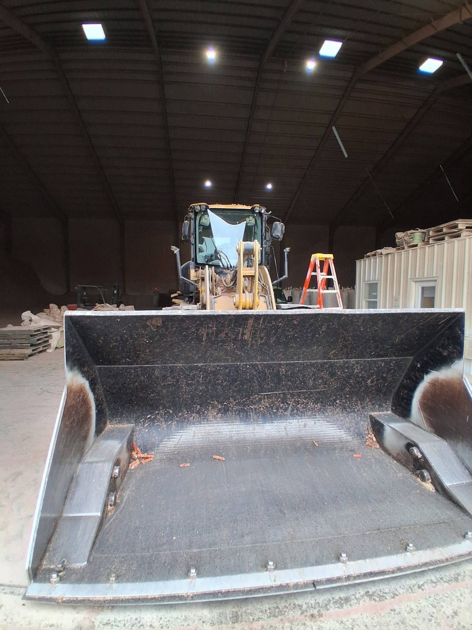 Front loader bucket in a dim warehouse, with machinery and an orange ladder in the background.