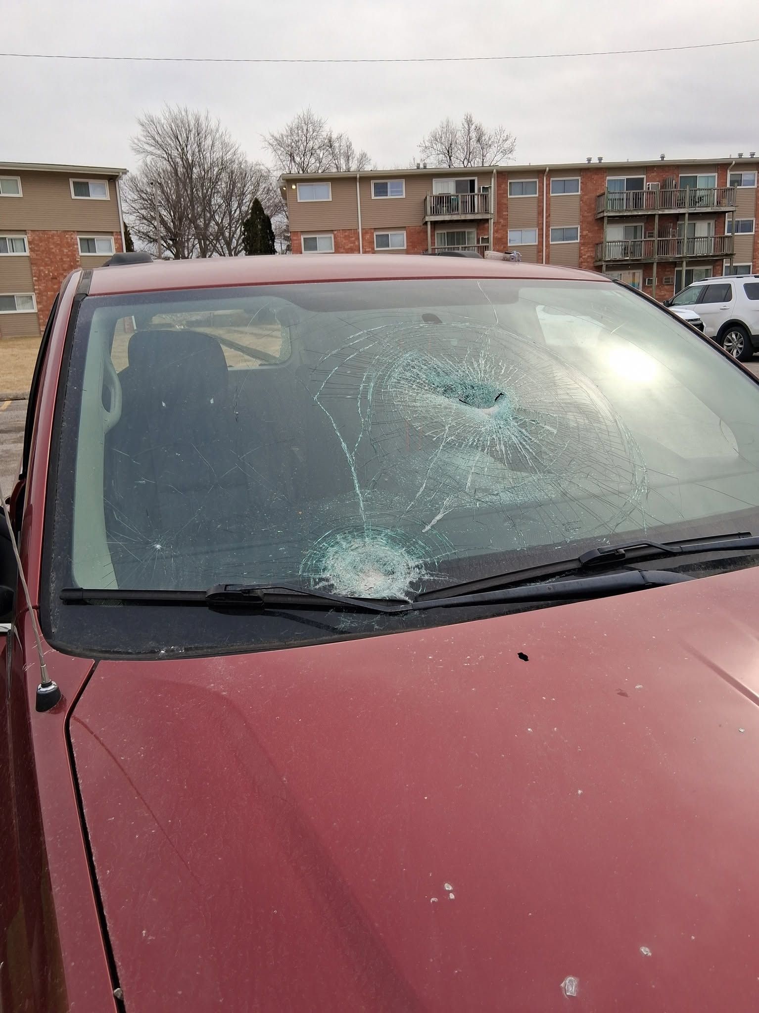 Red car windshield with a large crack and shattered impact area in a parking lot.