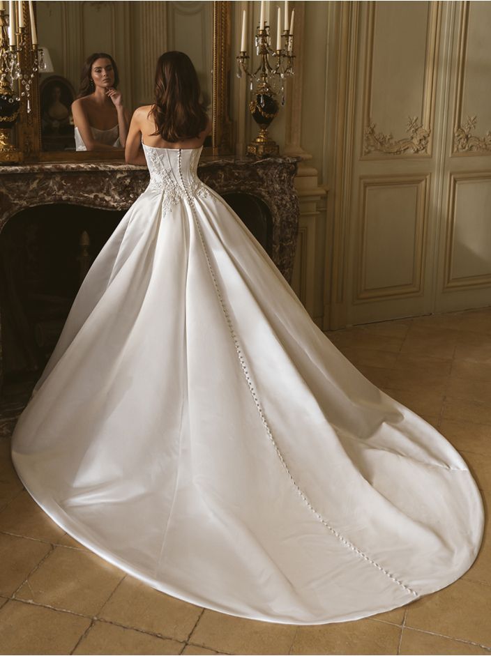 Braut im weißen trägerlosen Kleid, blickt in den Spiegel und steht in einem Raum mit kunstvollen Details.