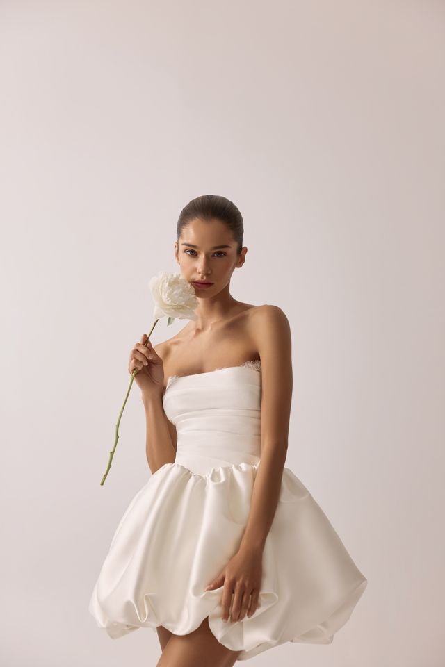 Frau in trägerlosem weißen Kleid, eine weiße Blume haltend, posiert vor einem schlichten Hintergrund.