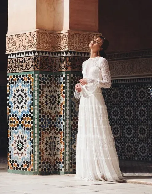 Eine Frau in einem weißen Kleid steht vor einer Mosaikwand.