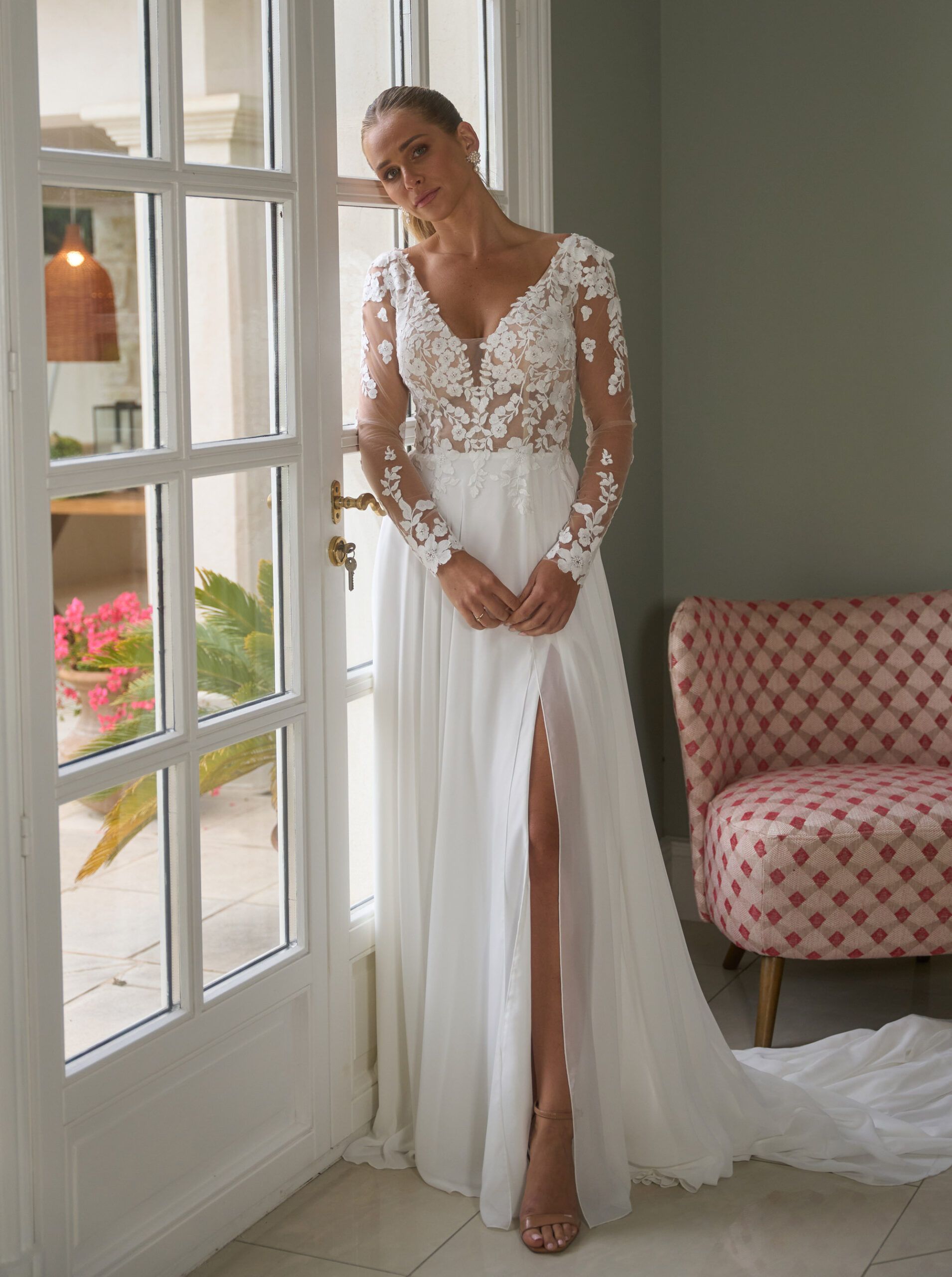 Eine Frau in einem weißen Brautkleid steht vor einem Fenster.