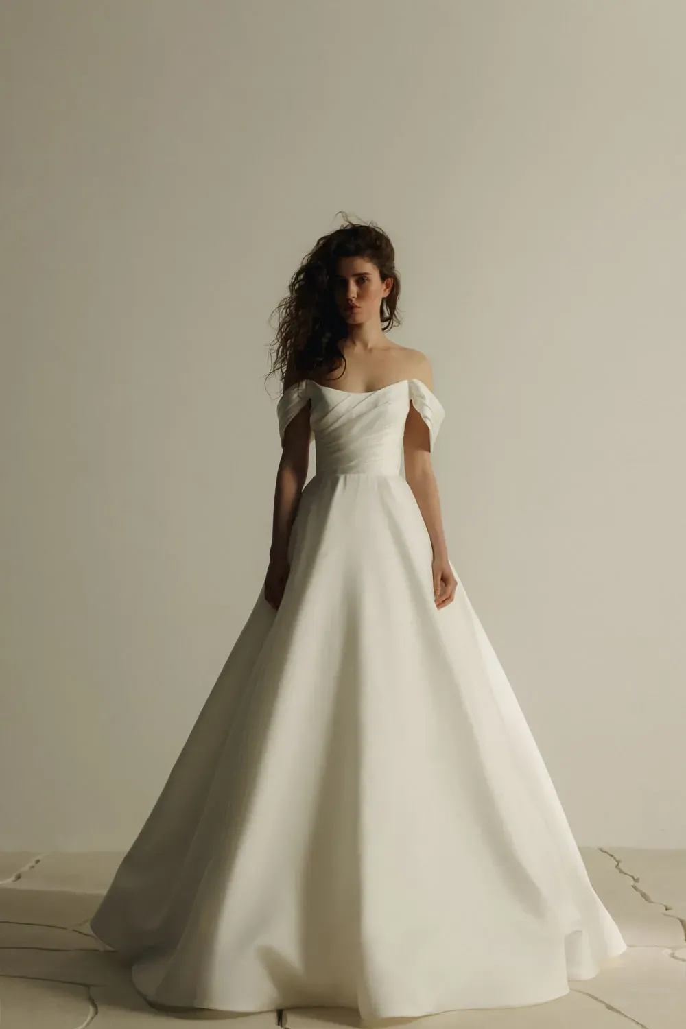 Frau in weißem schulterfreiem Brautkleid; steht vor einer weißen Wand.