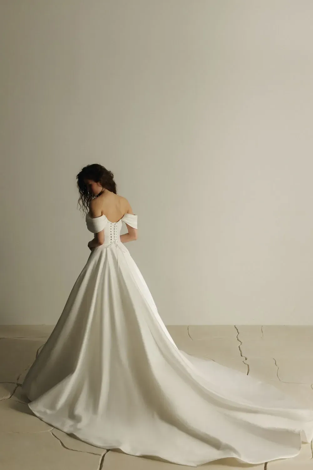 Frau in weißem schulterfreiem Brautkleid, mit dem Rücken zur Kamera, posiert auf rissigem Boden.