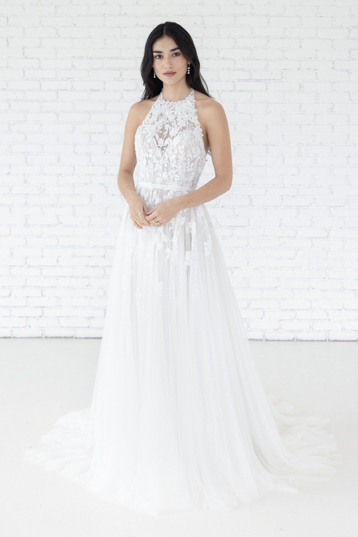 Eine Frau in einem weißen Brautkleid steht vor einer weißen Backsteinmauer.