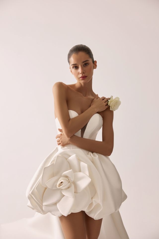 Frau in einem weißen trägerlosen Kleid mit einem großen Blumenakzent, hält eine Rose. Weißer Hintergrund.