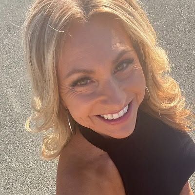Smiling woman in a black sleeveless top, taking a selfie outdoors in bright sunlight.