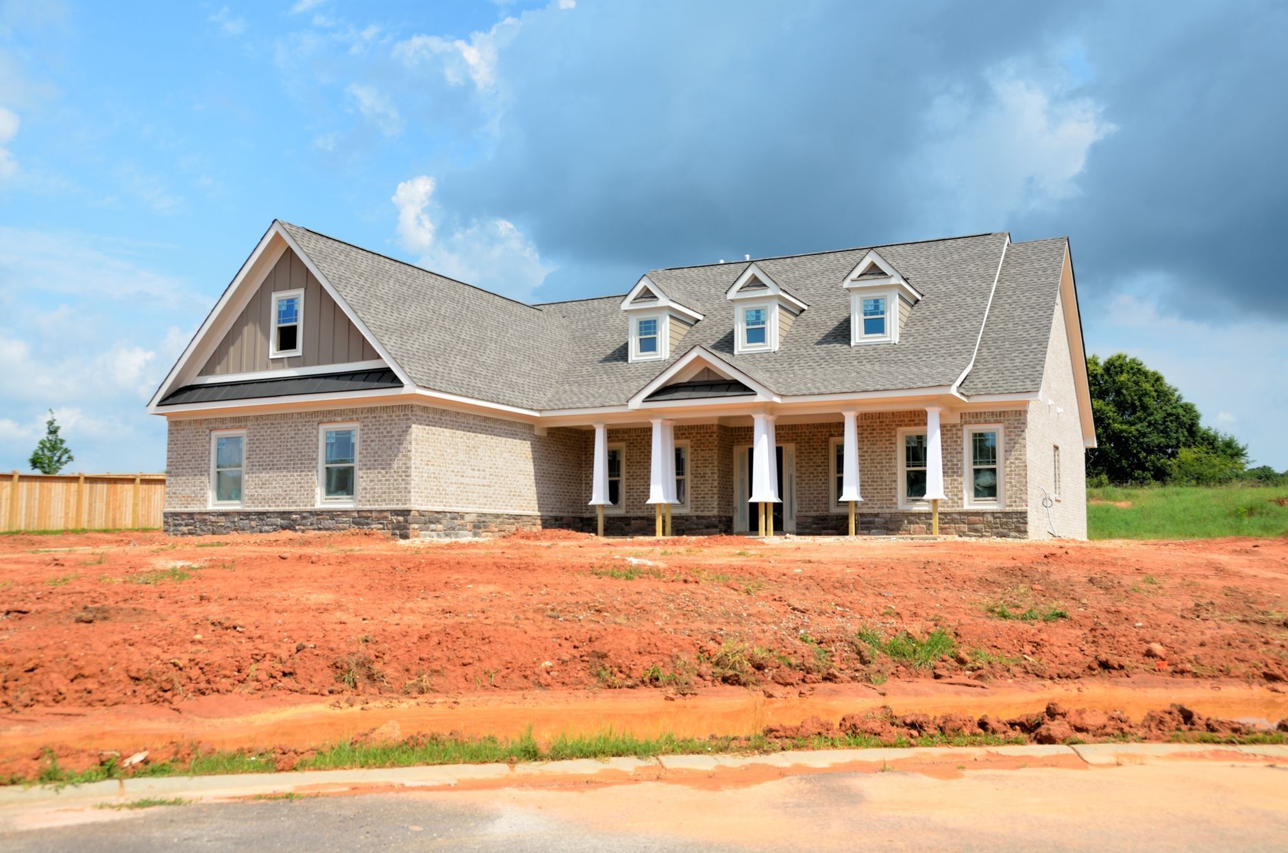 Large gray suburban house under construction with a red dirt front yard and blue sky