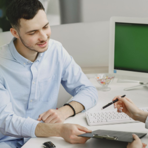 Man in light blue shirt signing paperwork across a desk from another person with a computer monitor nearby