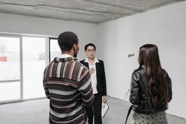Three people stand in an empty white room, talking near large windows.