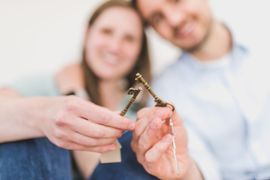 Hands holding a small house key and keychain, with a smiling couple blurred in the background.