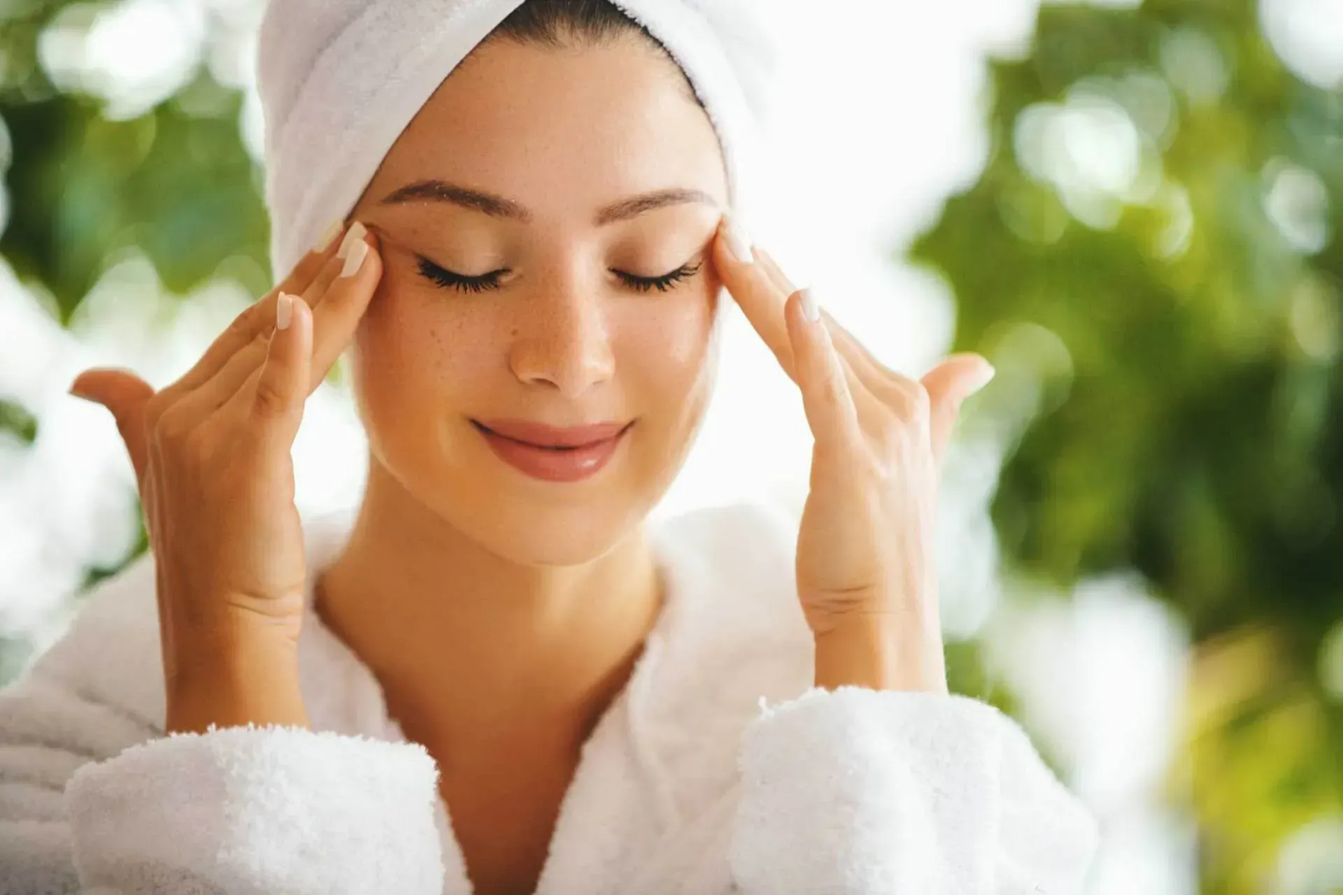 Woman in a white robe and towel, eyes closed, gently massages her eye area. Green foliage in background.