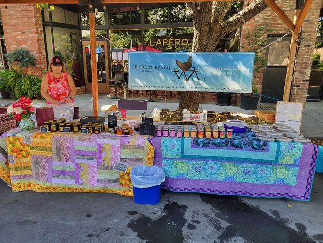 A vendor stands behind two tables covered in patterned quilts, displaying jars of jam and goods at an outdoor market.