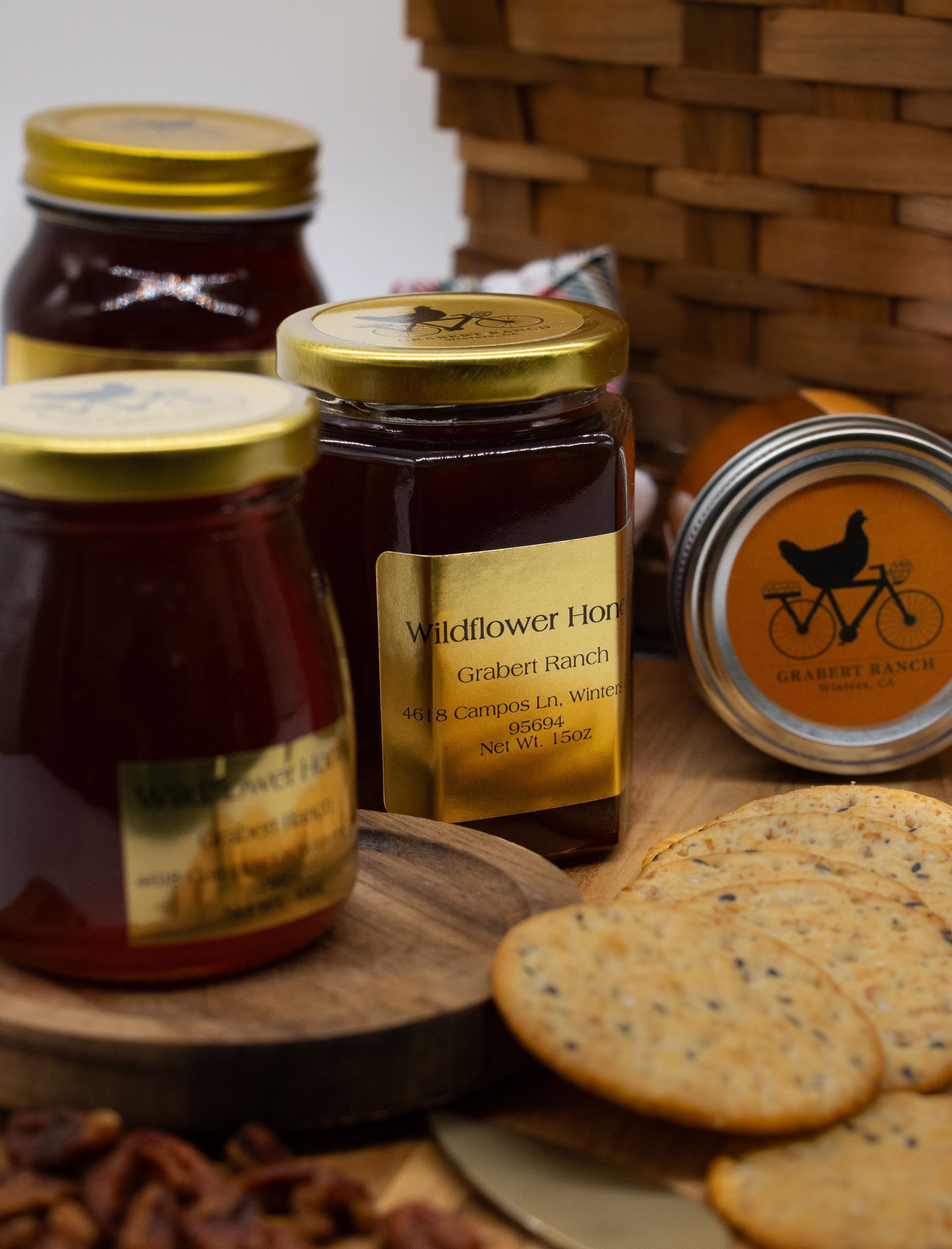 Three jars of preserves and a stack of seeded crackers on a wooden surface with a wicker basket in the background.