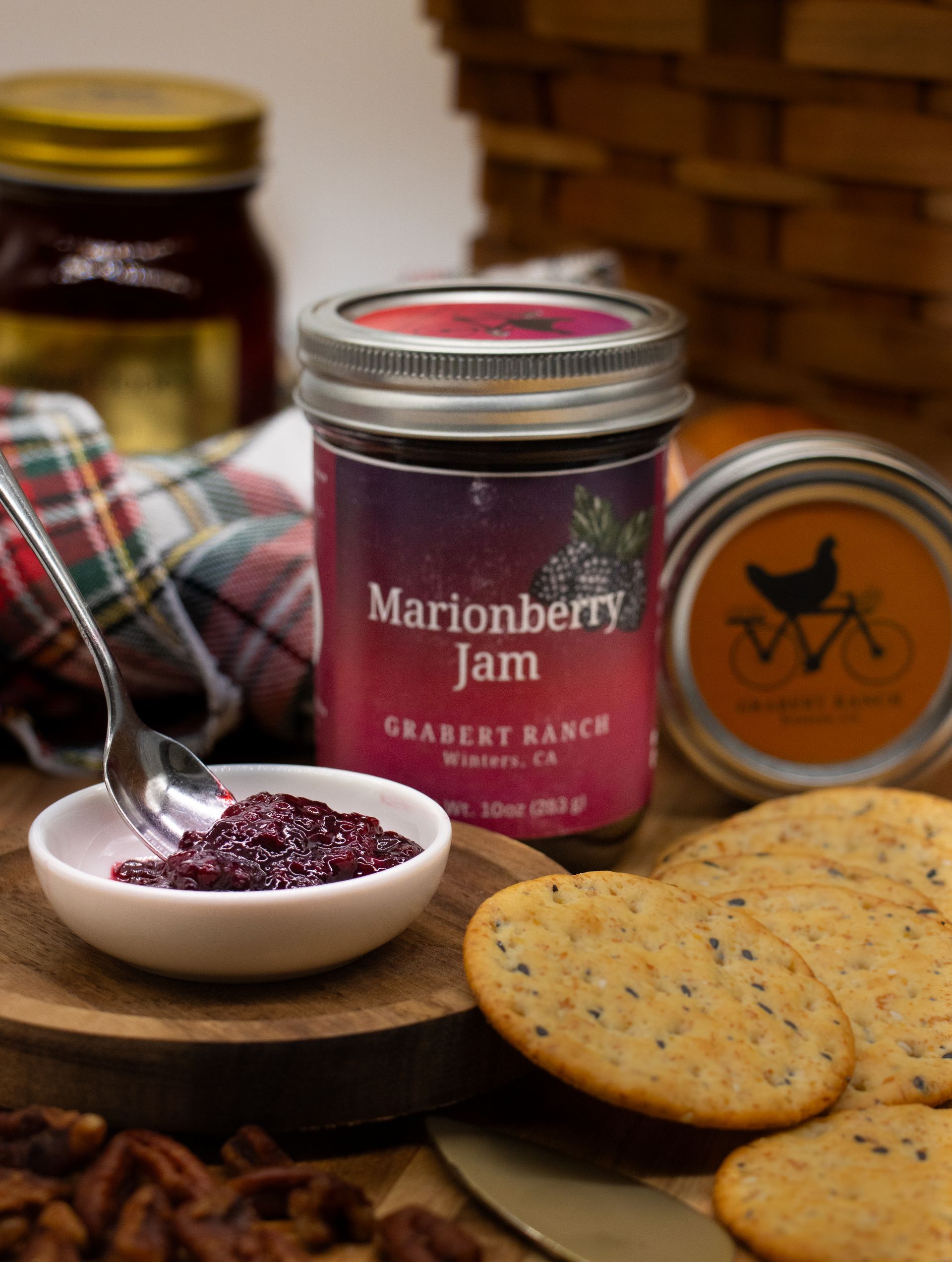 A jar of Marionberry Jam, a small bowl of jam with a spoon, crackers, and pecans on a wooden surface.