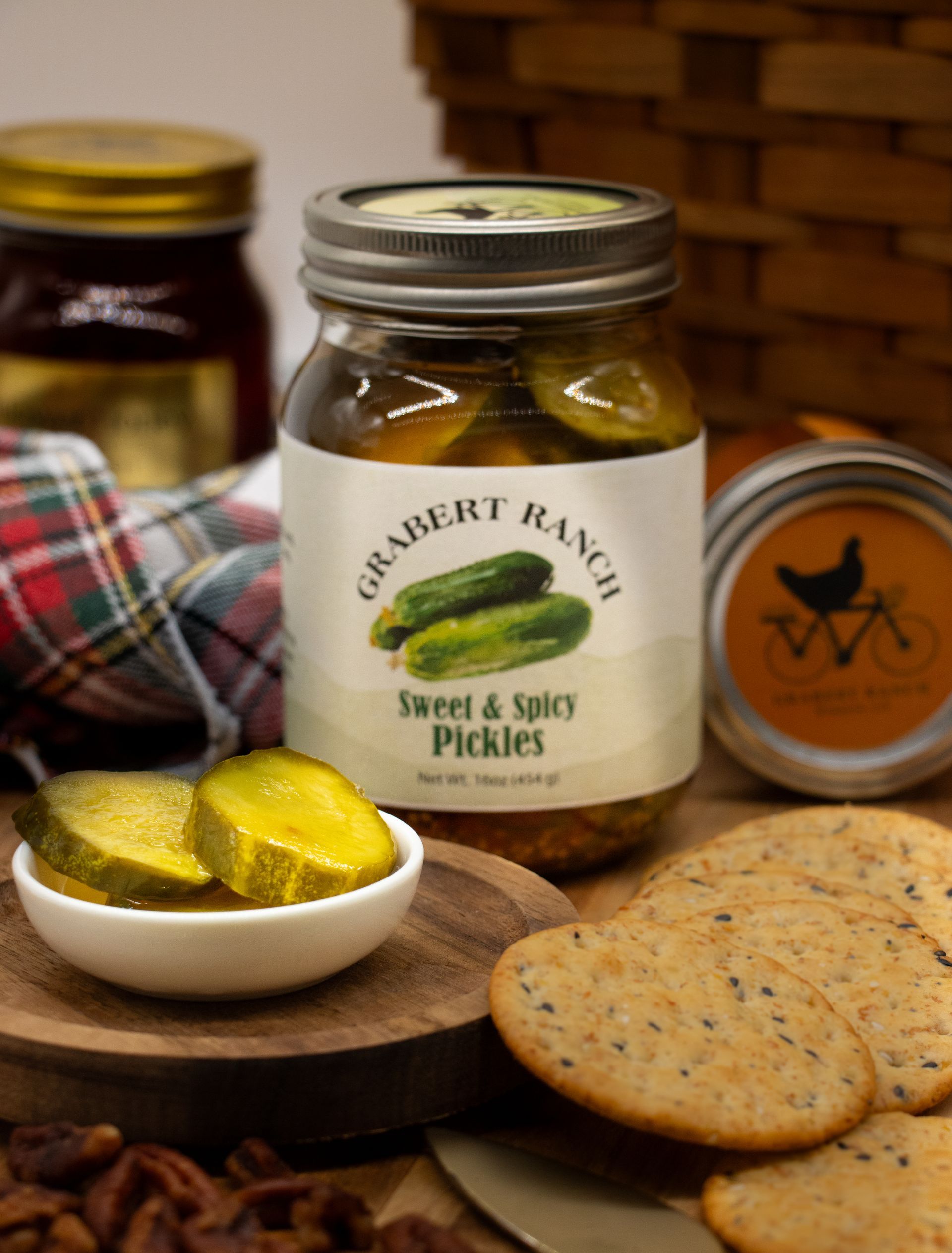 A jar of Grabert Ranch Sweet & Spicy Pickles with a small dish of pickle slices, crackers, and nuts on a wooden surface.