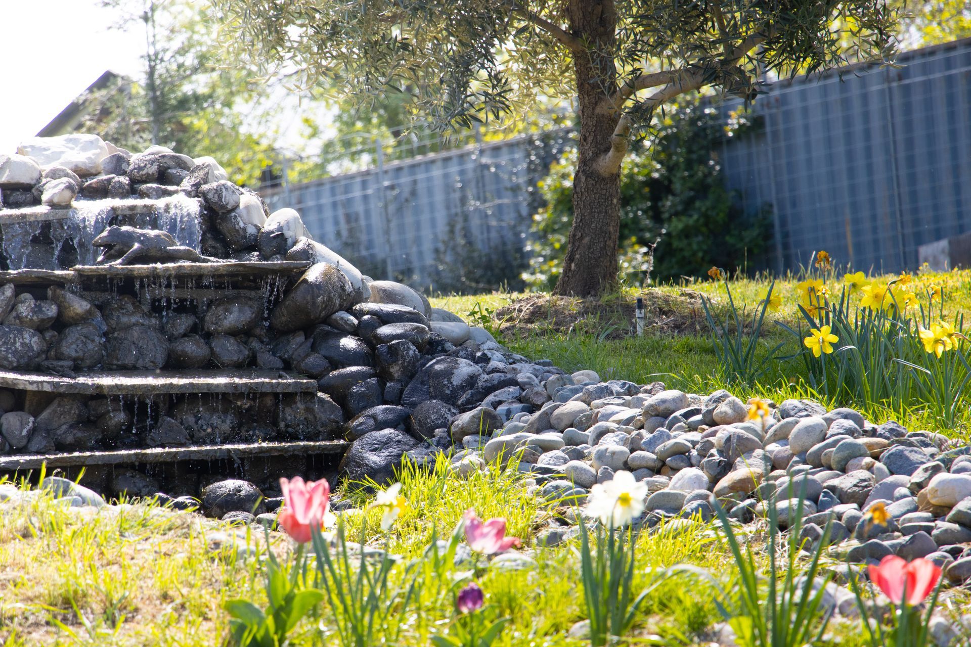 A tiered stone waterfall flows into a rocky stream, surrounded by colorful spring tulips and daffodils in a garden.