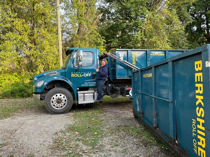 Worker beside teal dump truck and blue container in wooded gravel lot
