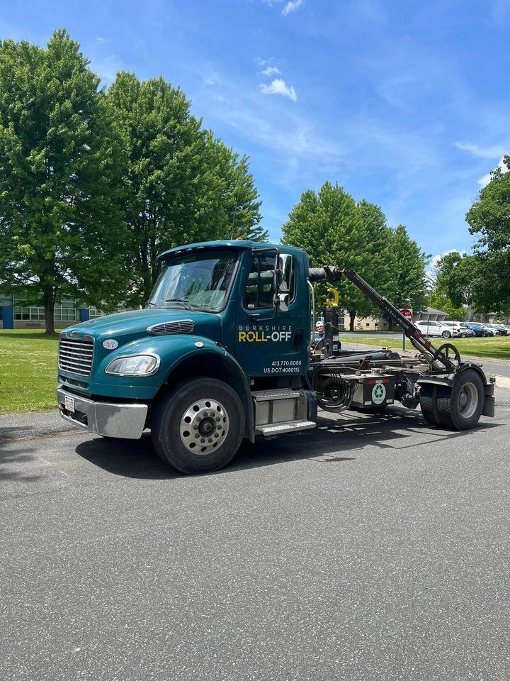 Teal tow truck parked on a road beside green trees under a blue sky.
