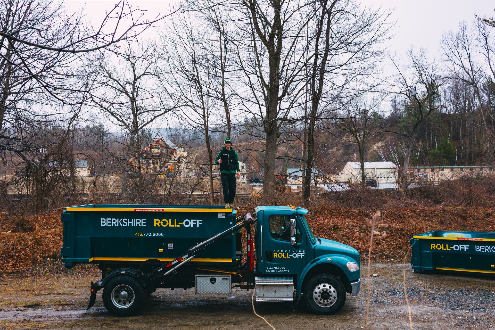 Green dump truck with “ROLL OFF” parked on a roadside beside bare trees in a muddy field
