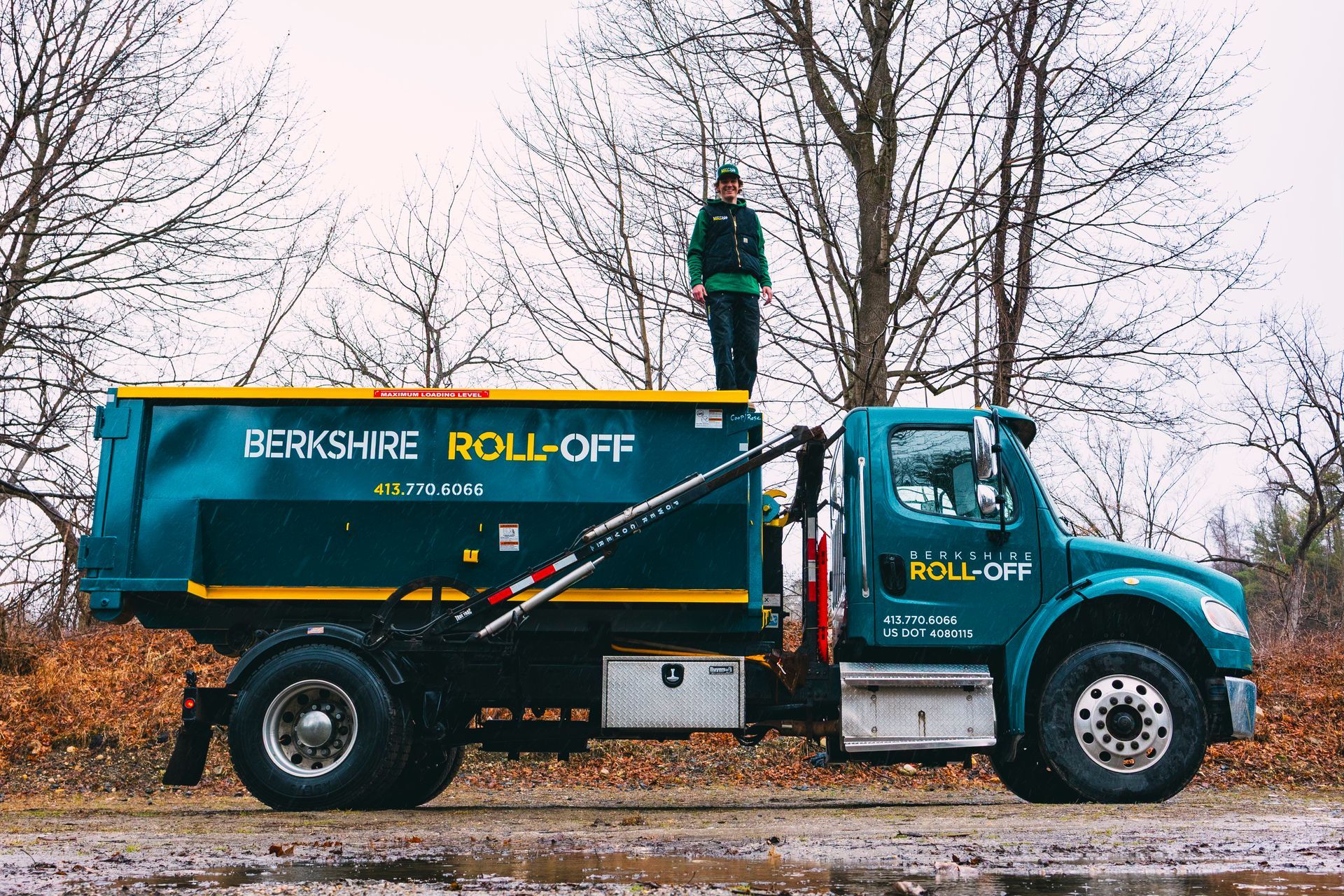Blue Berkshire Roll-Off dump truck with a person standing on top, parked by leafless trees.