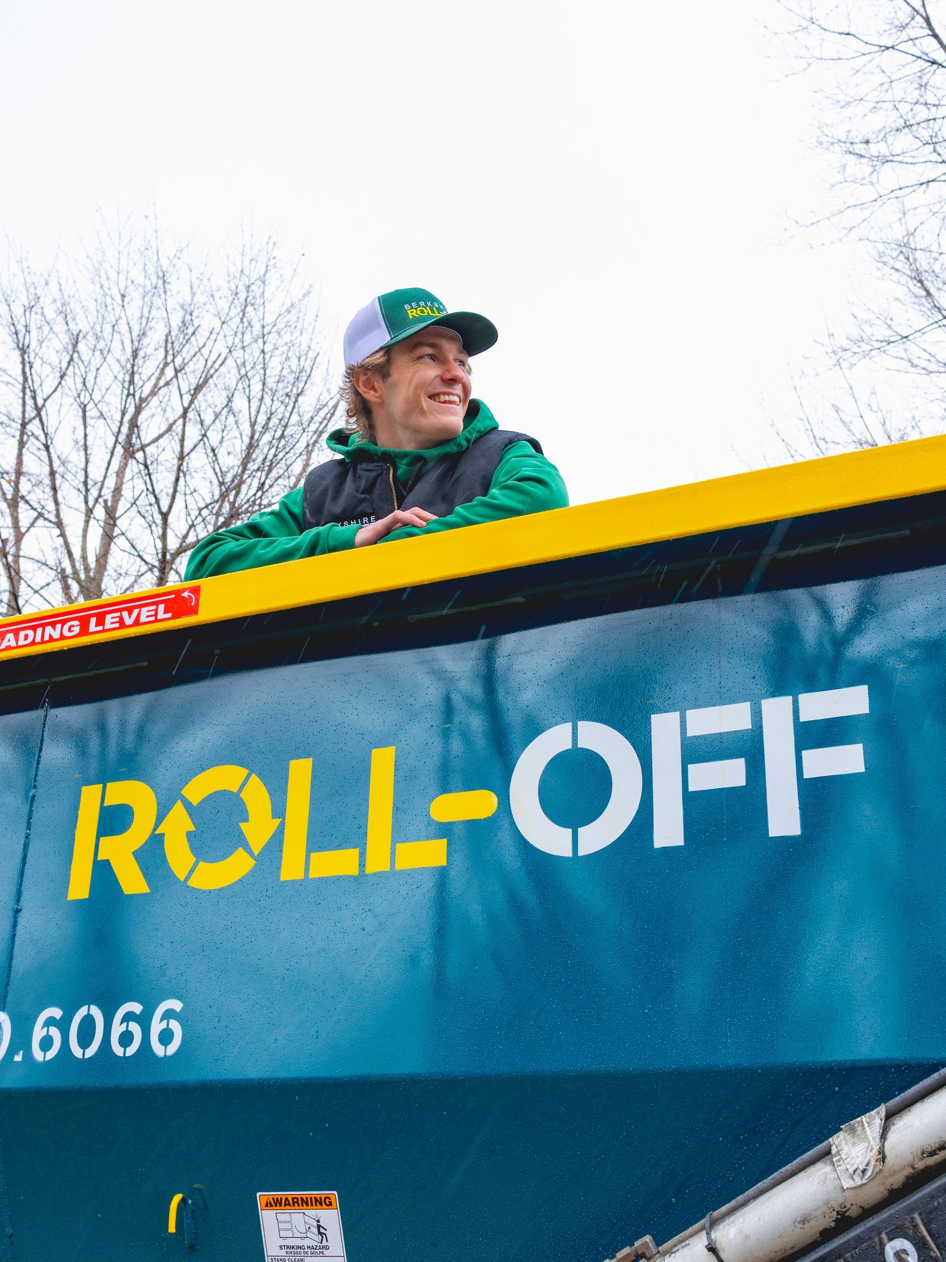 Person in green jacket and cap standing in a blue roll-off dumpster, smiling outdoors