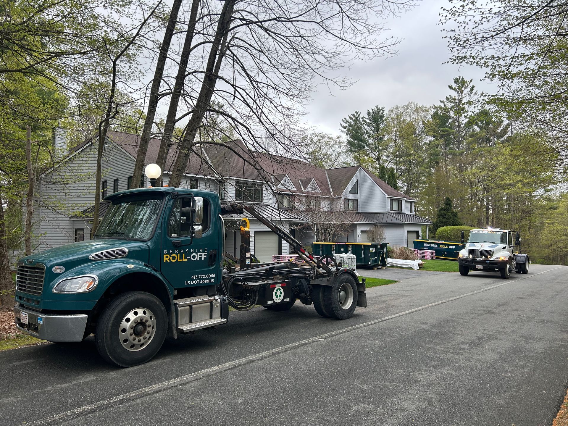 Tow truck parked on a residential street beside houses and trees, with another truck in the background
