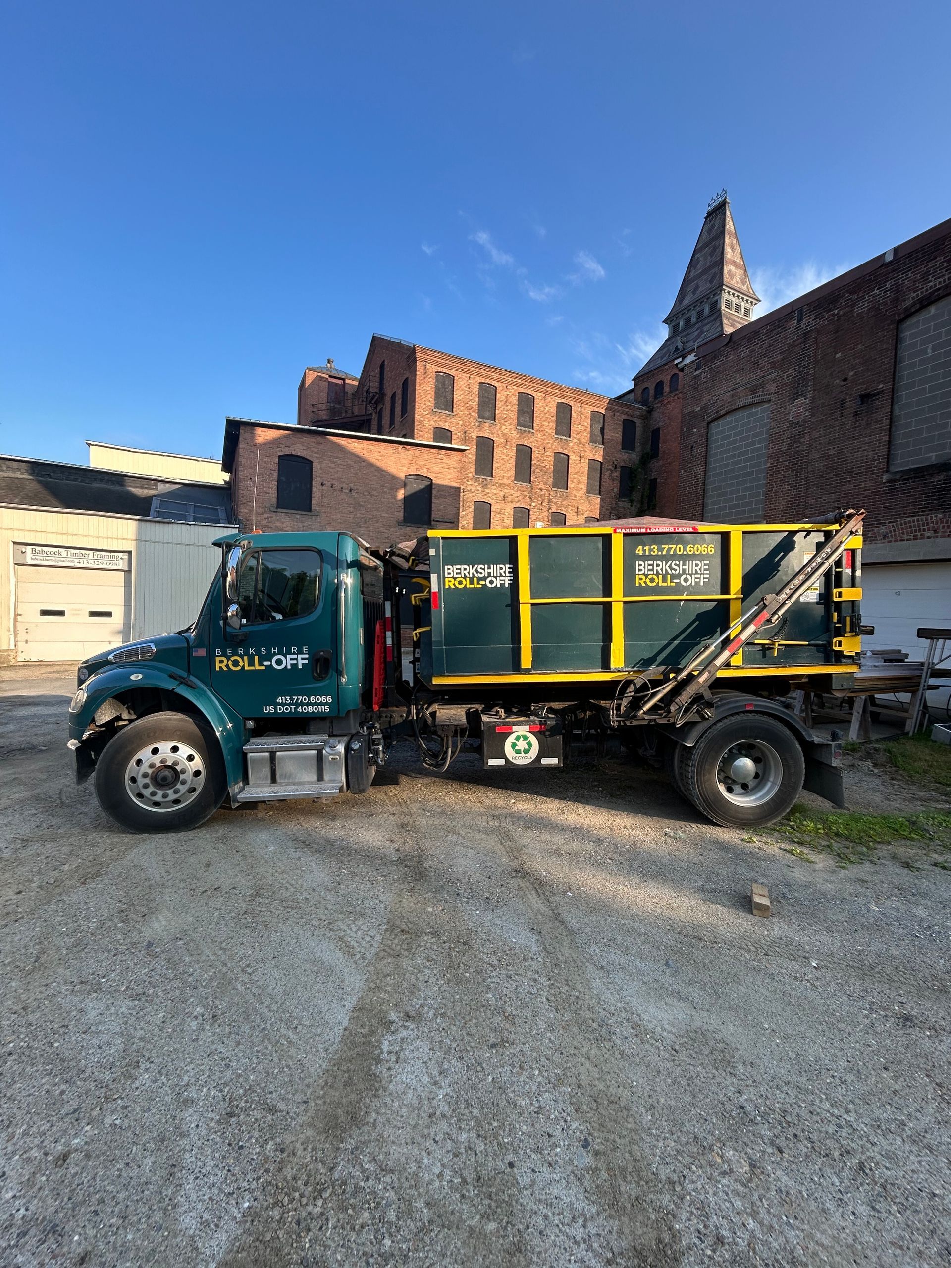 Green and yellow dump truck parked on gravel in front of brick buildings at dusk