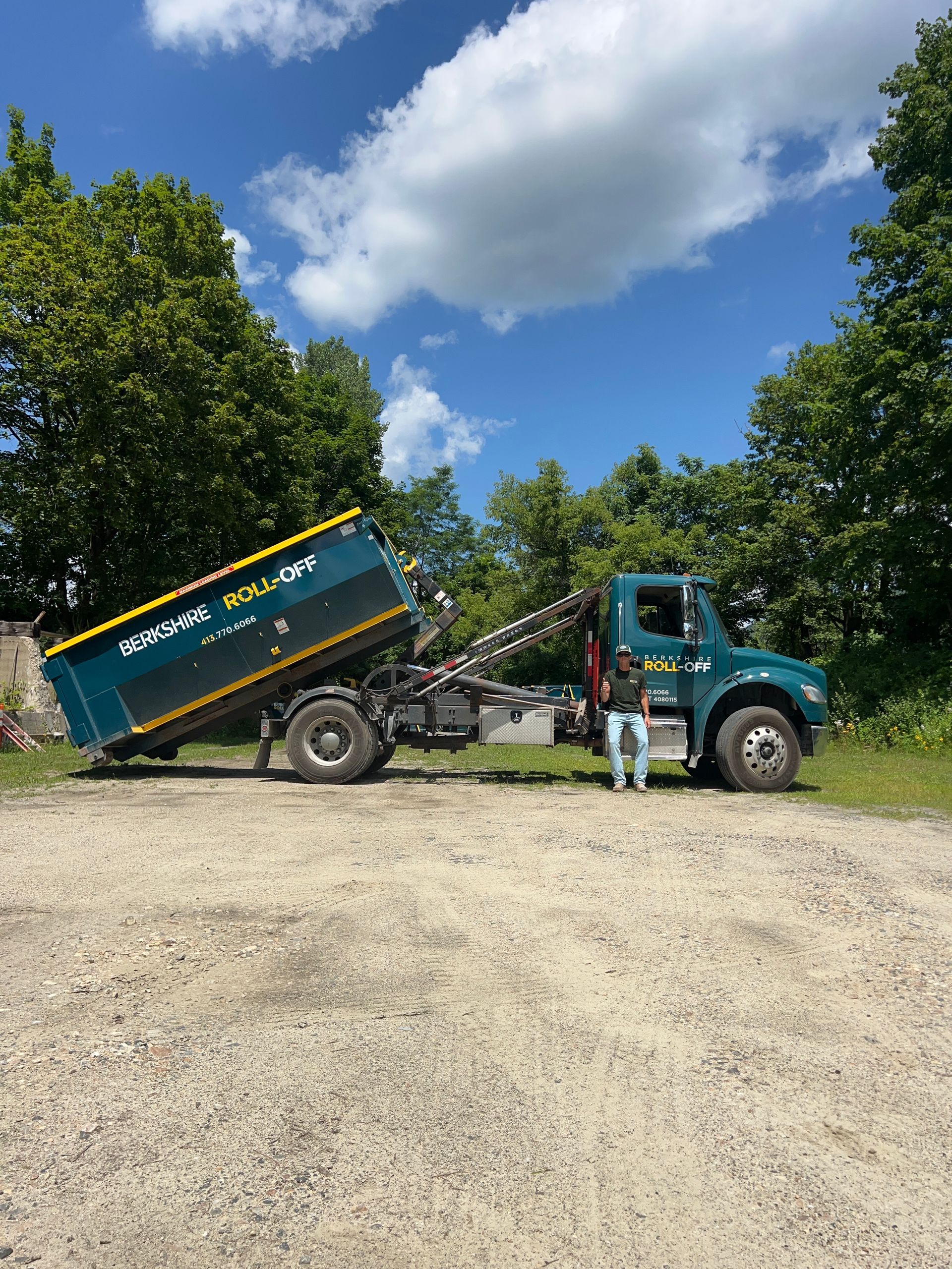 Dump truck parked on a gravel road with a person standing beside it under a blue sky with trees.