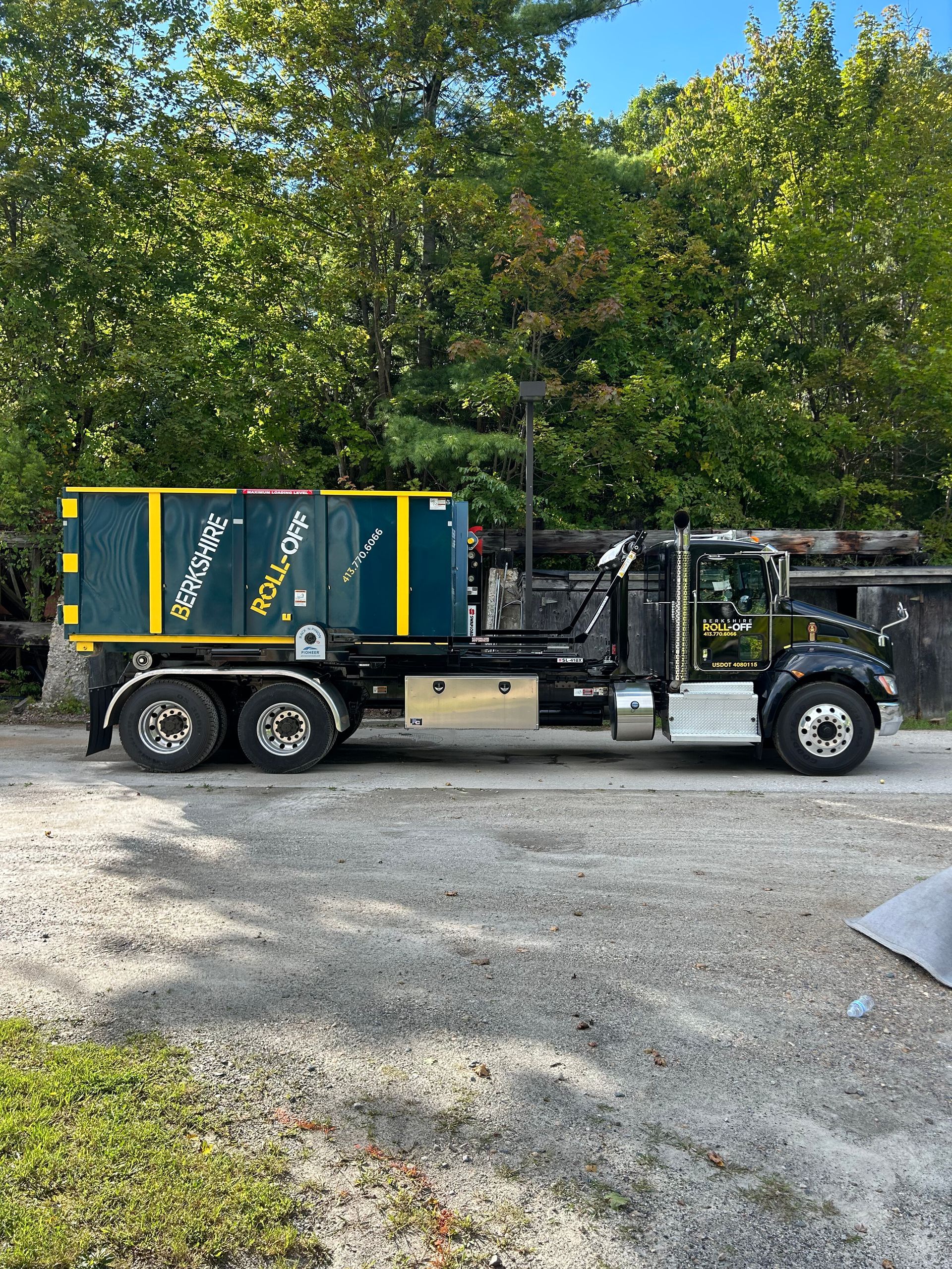Black utility truck with blue-green cargo bed and yellow trim parked on gravel under trees.