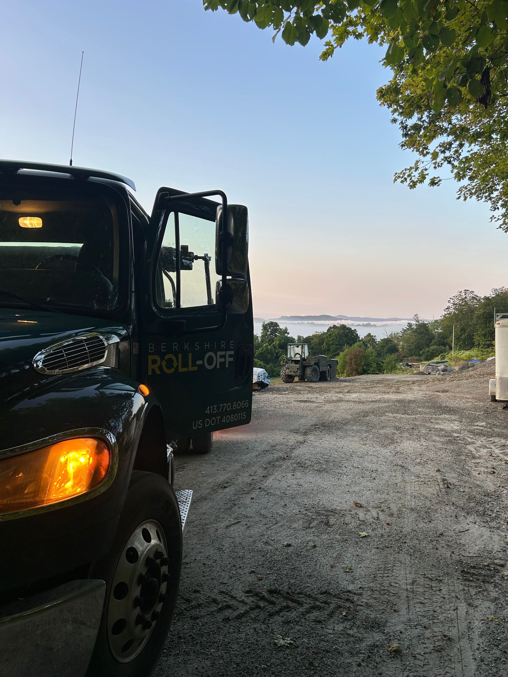 Black dump truck on gravel driveway at sunset overlooking water, with trees and a clear sky.