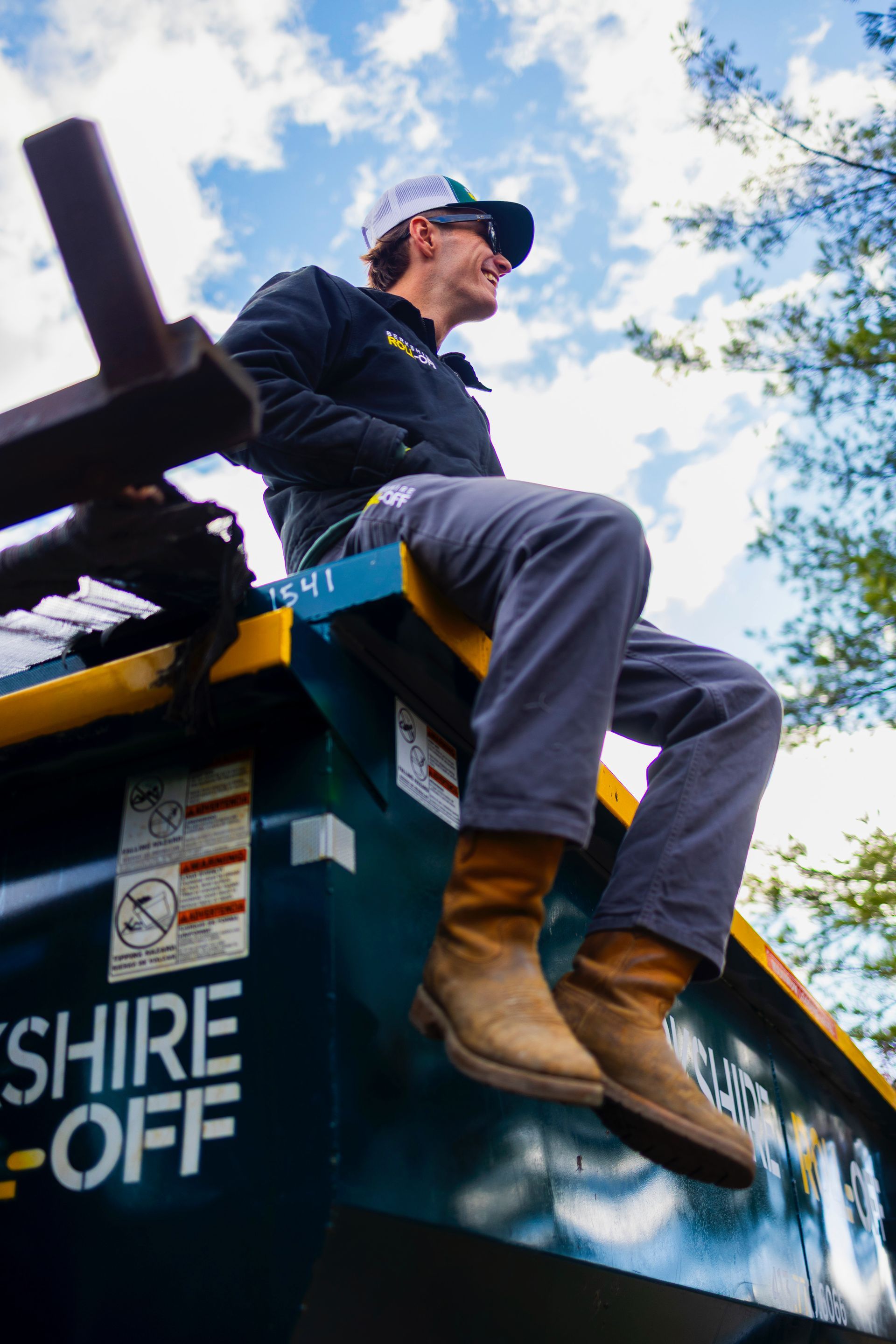 Person sitting on a large green dumpster outdoors, wearing a blue cap, black jacket, jeans, and tan boots.