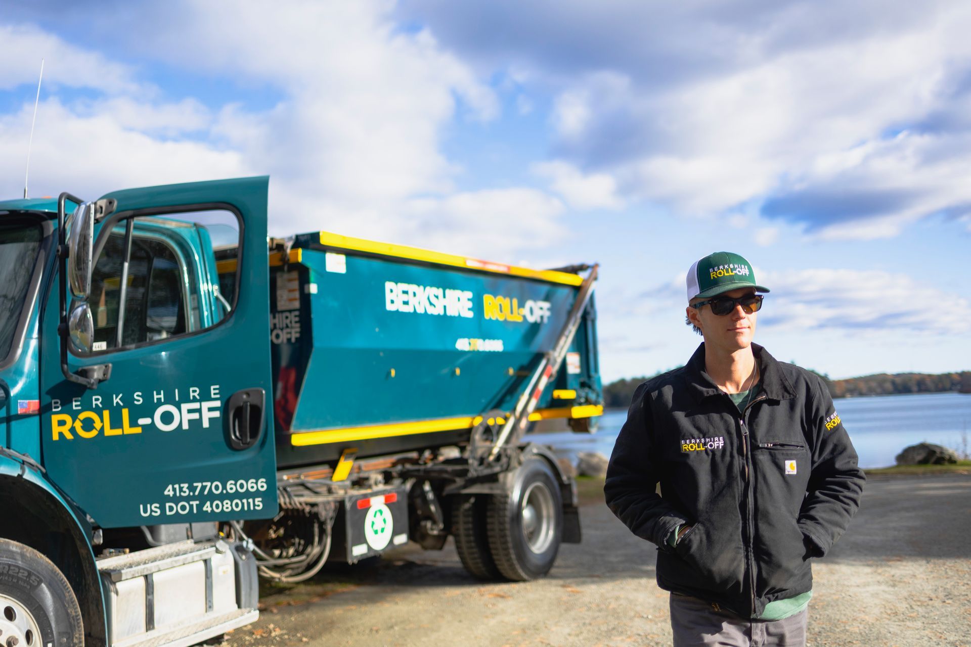 Worker stands beside a teal and yellow roll-off truck by the water under a cloudy sky.