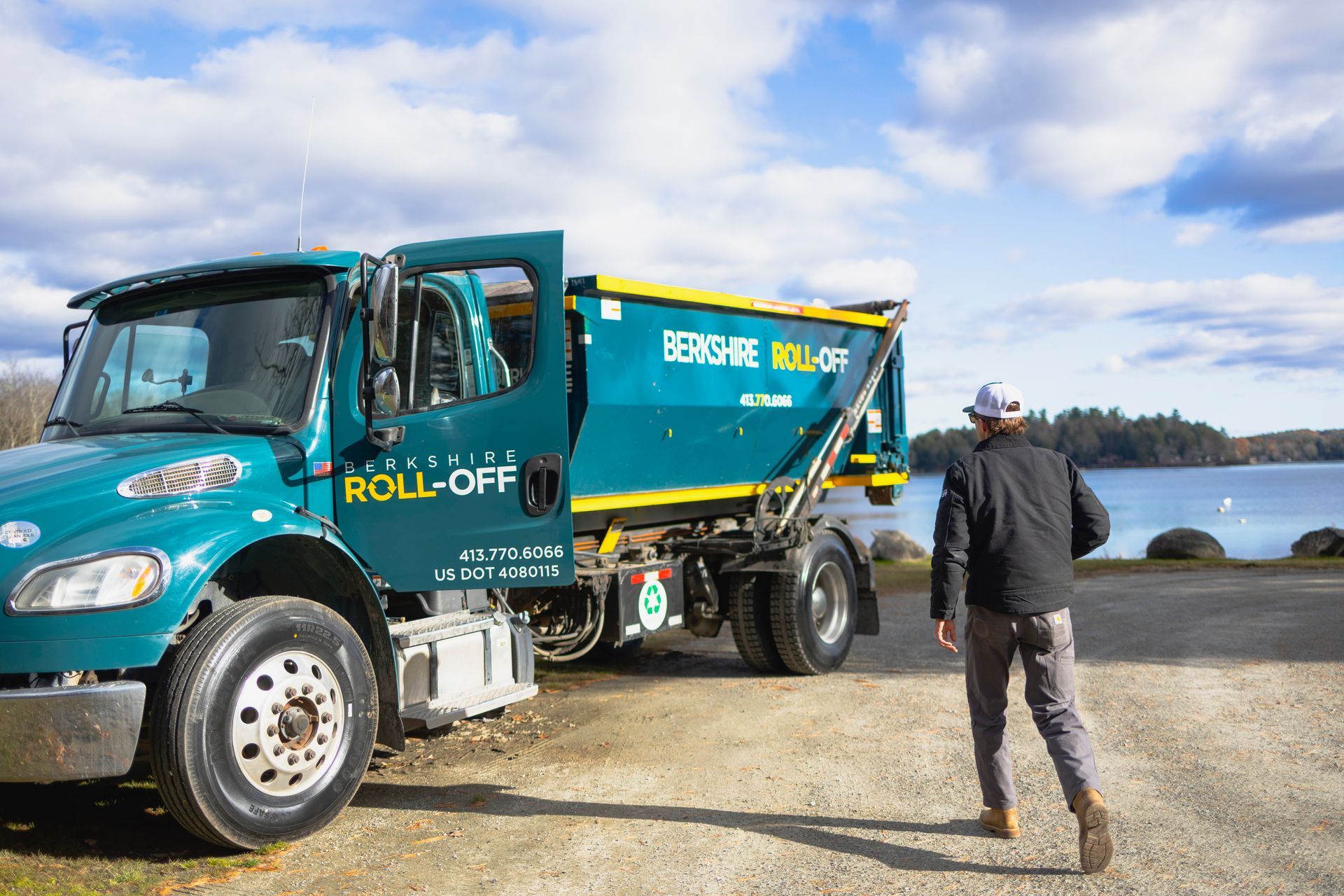 Turquoise and yellow roll-off truck by the water, with a person standing nearby on a gravel lot.