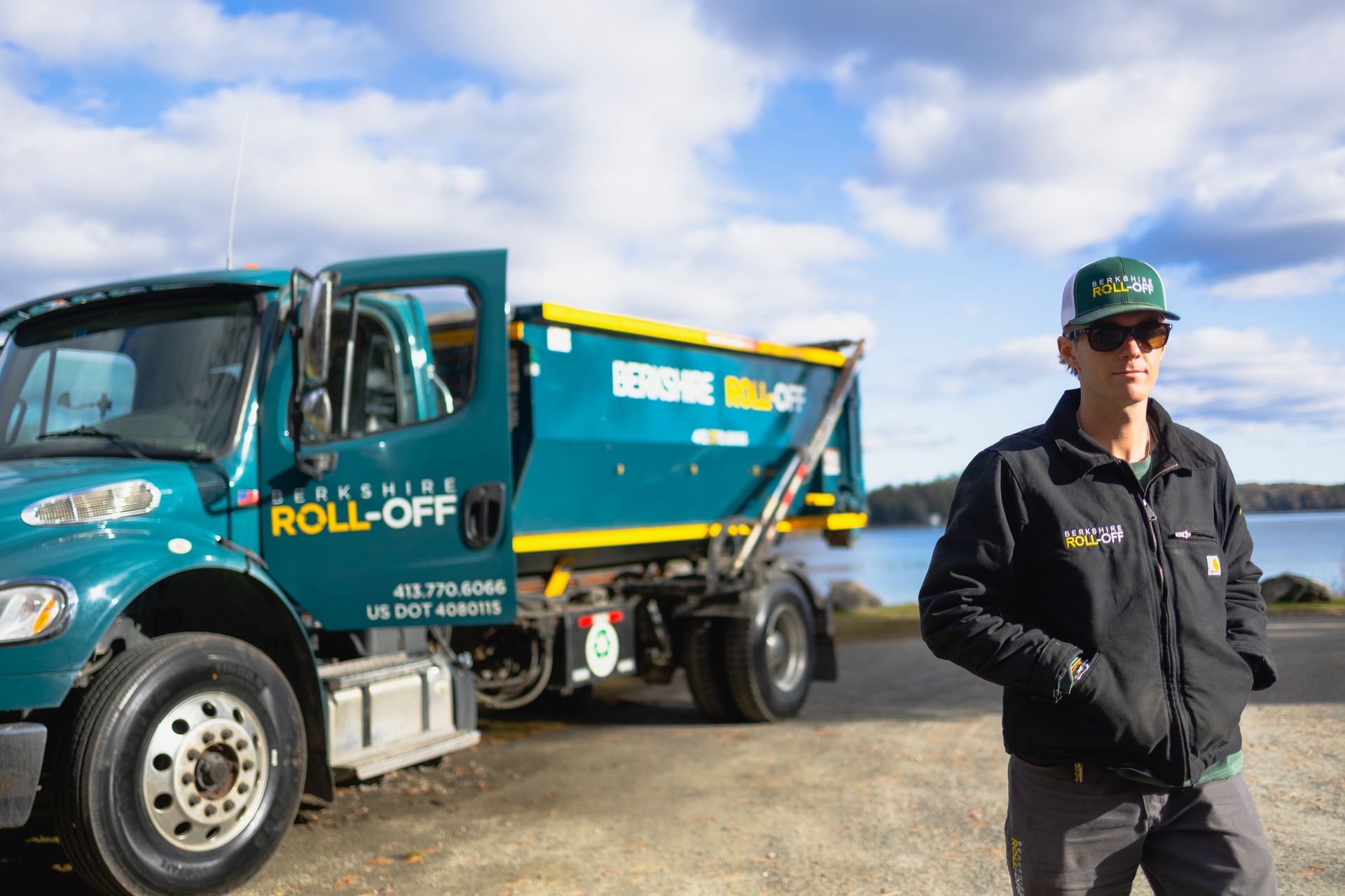 Teal roll-off truck parked beside a worker in a black jacket and cap on a gravel lot.