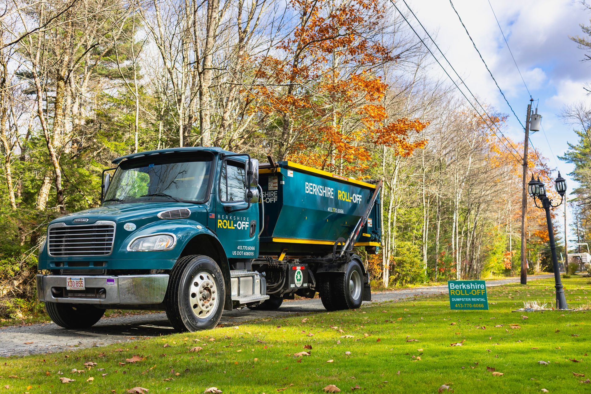 Blue dump truck parked on a grassy roadside with autumn trees and power lines in the background