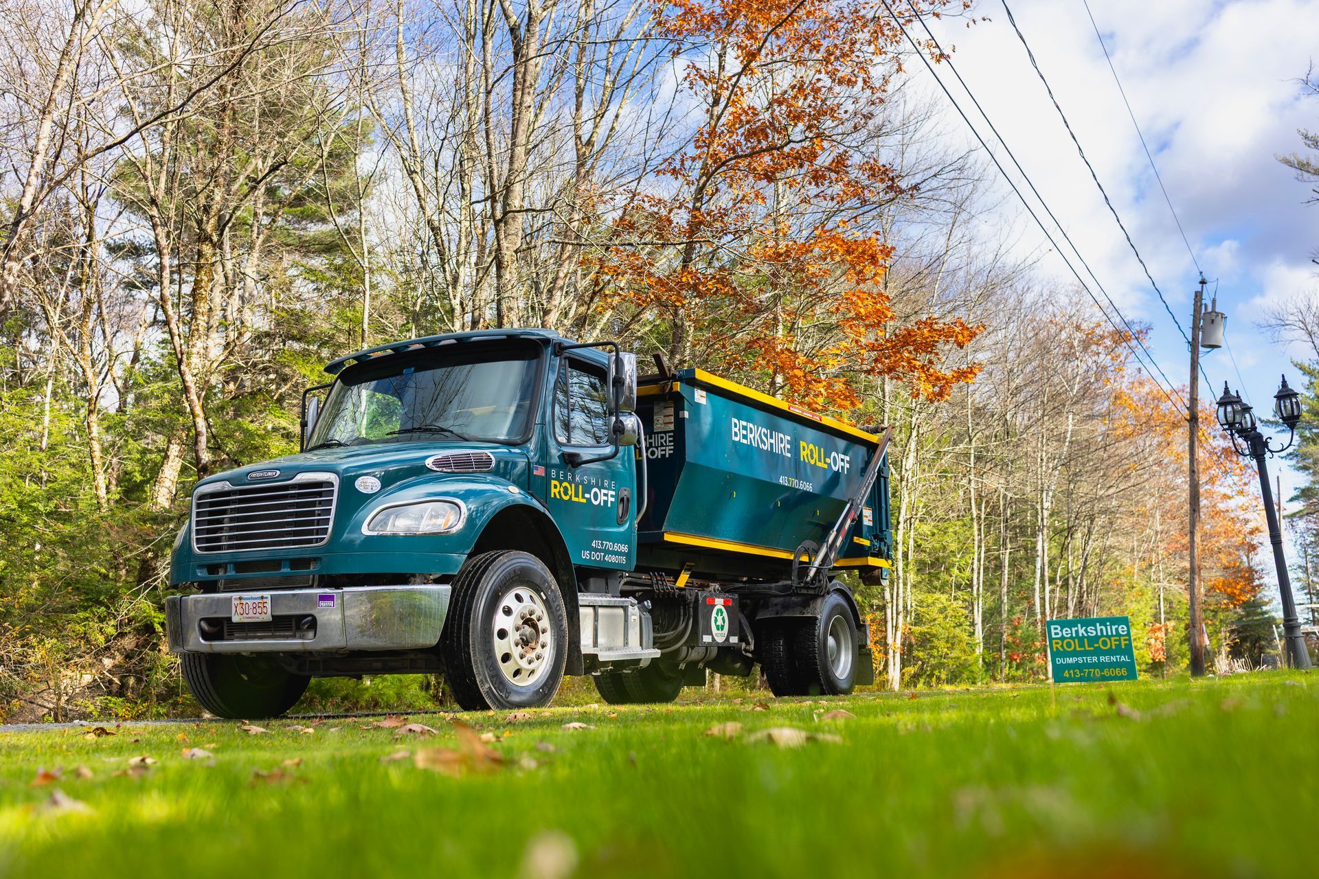 Green dump truck on a grassy roadside in autumn trees under a blue sky.