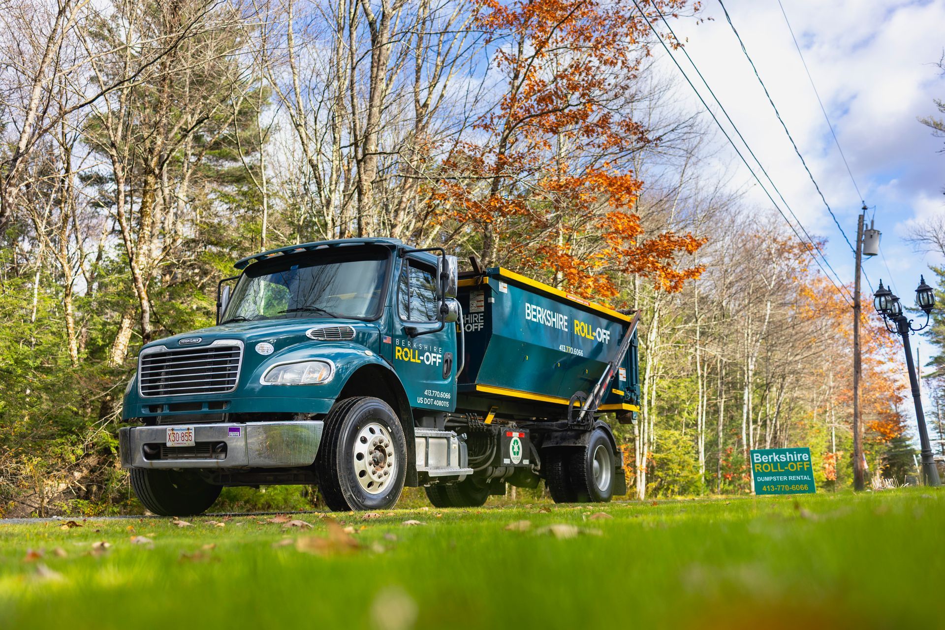 Teal dump truck driving on a rural road beside autumn trees under a partly cloudy sky