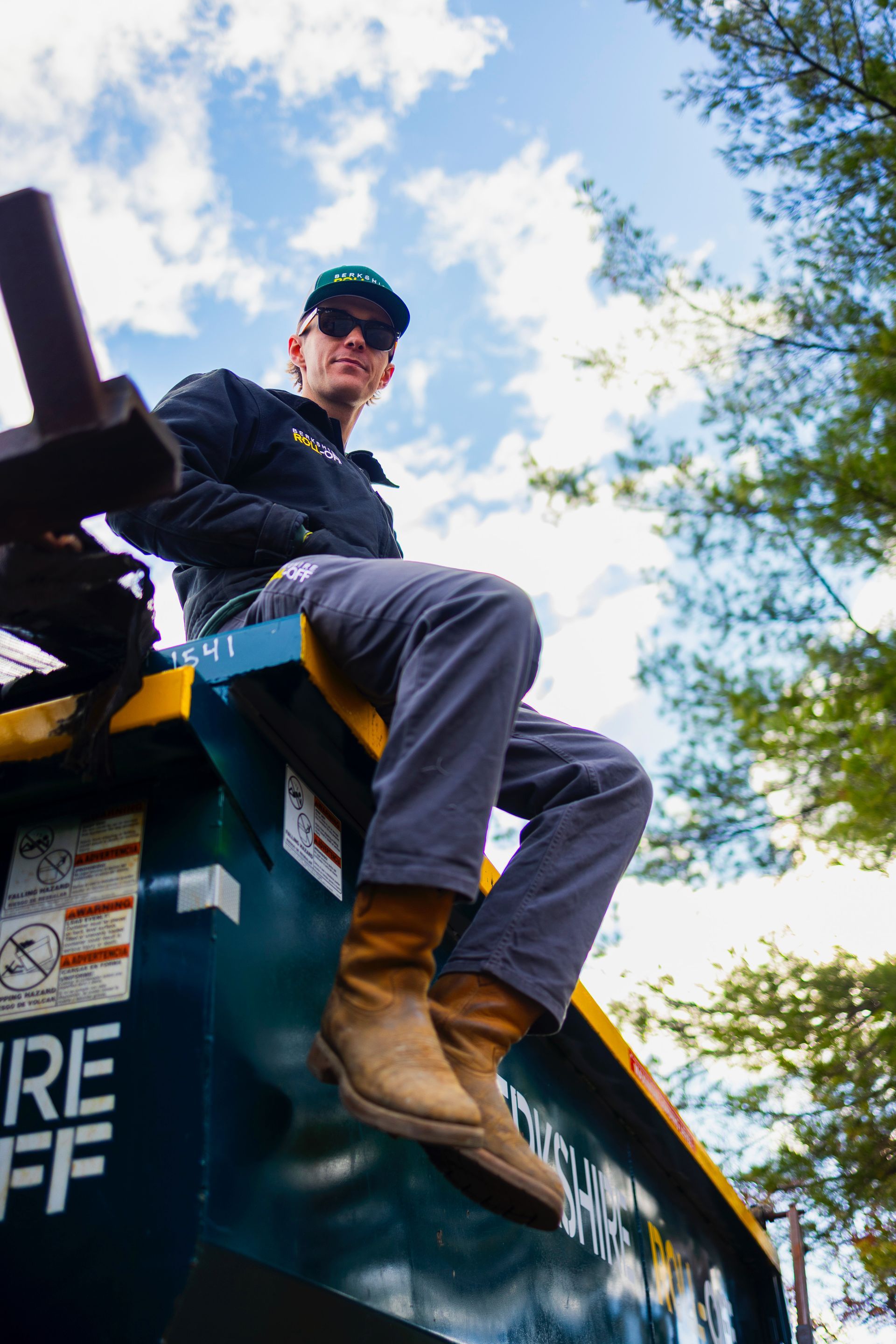 Worker sitting on top of a dumpster, wearing a cap and work clothes under a blue sky