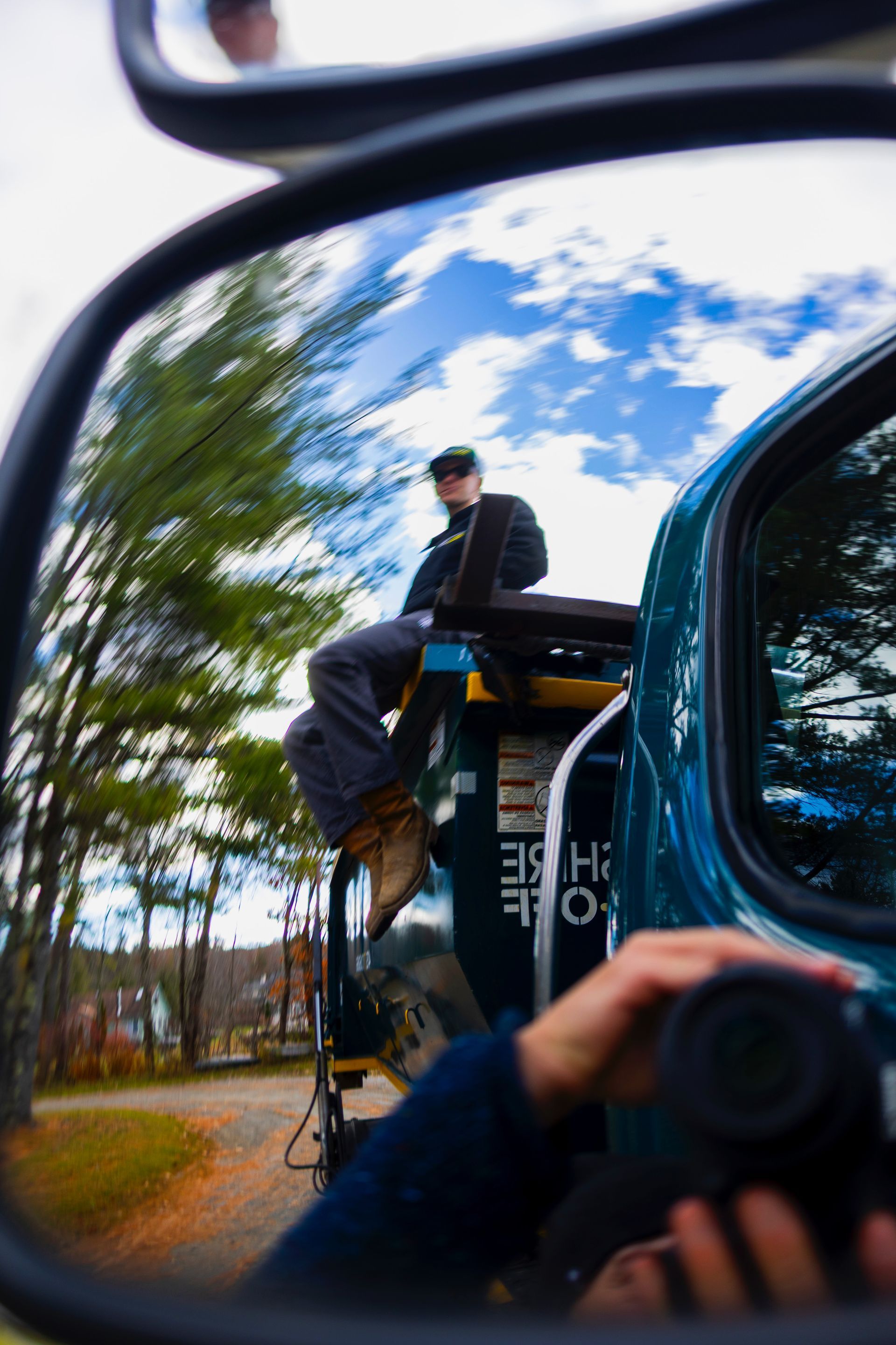 Person riding on the back of a truck, seen in a side mirror, with trees and blue sky reflected.