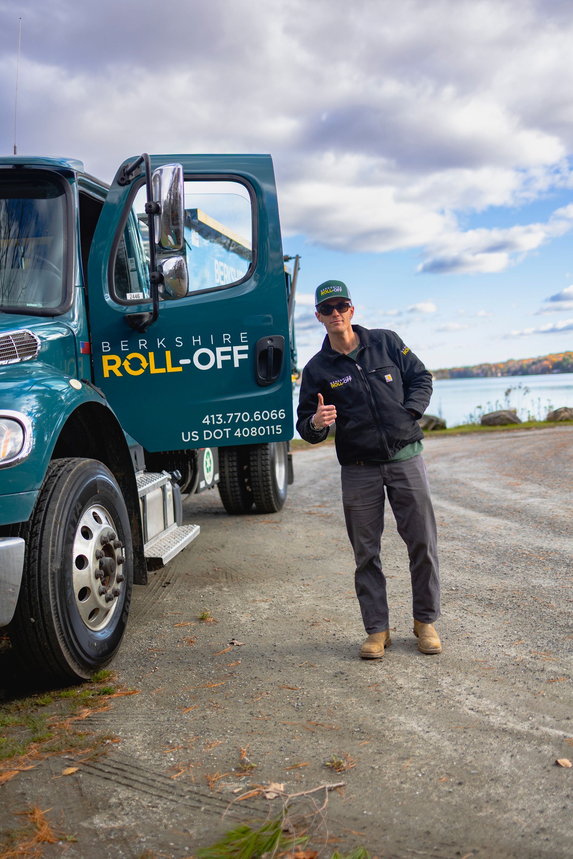 Worker standing beside a teal roll-off truck near the water under a cloudy sky