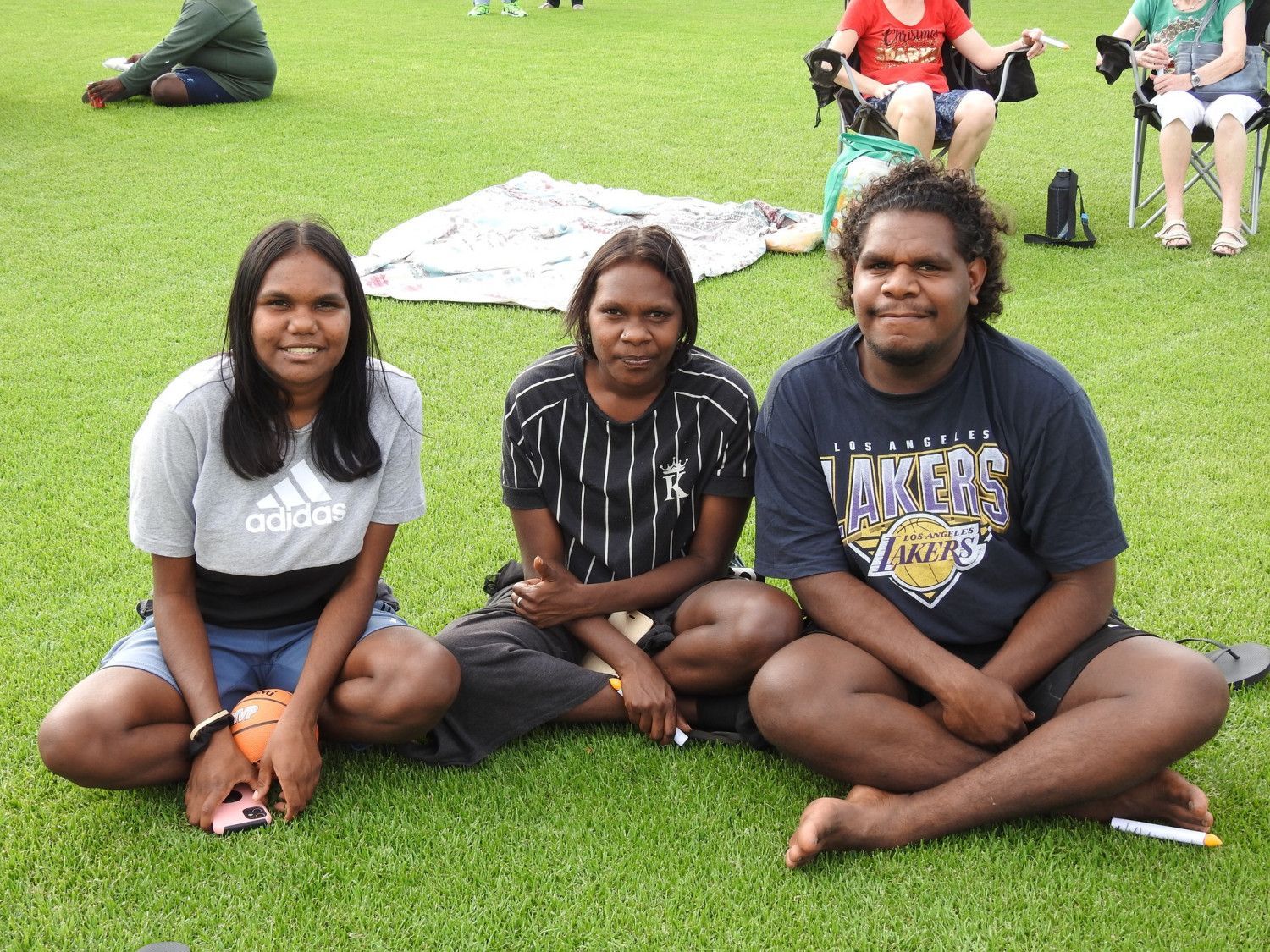 Three young people sit on grass, smiling. One in a Lakers shirt, one in a striped shirt, one in an Adidas top.