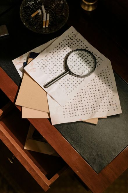 A magnifying glass rests on a paper filled with small text atop a dark wooden table with an ashtray containing cigarettes.
